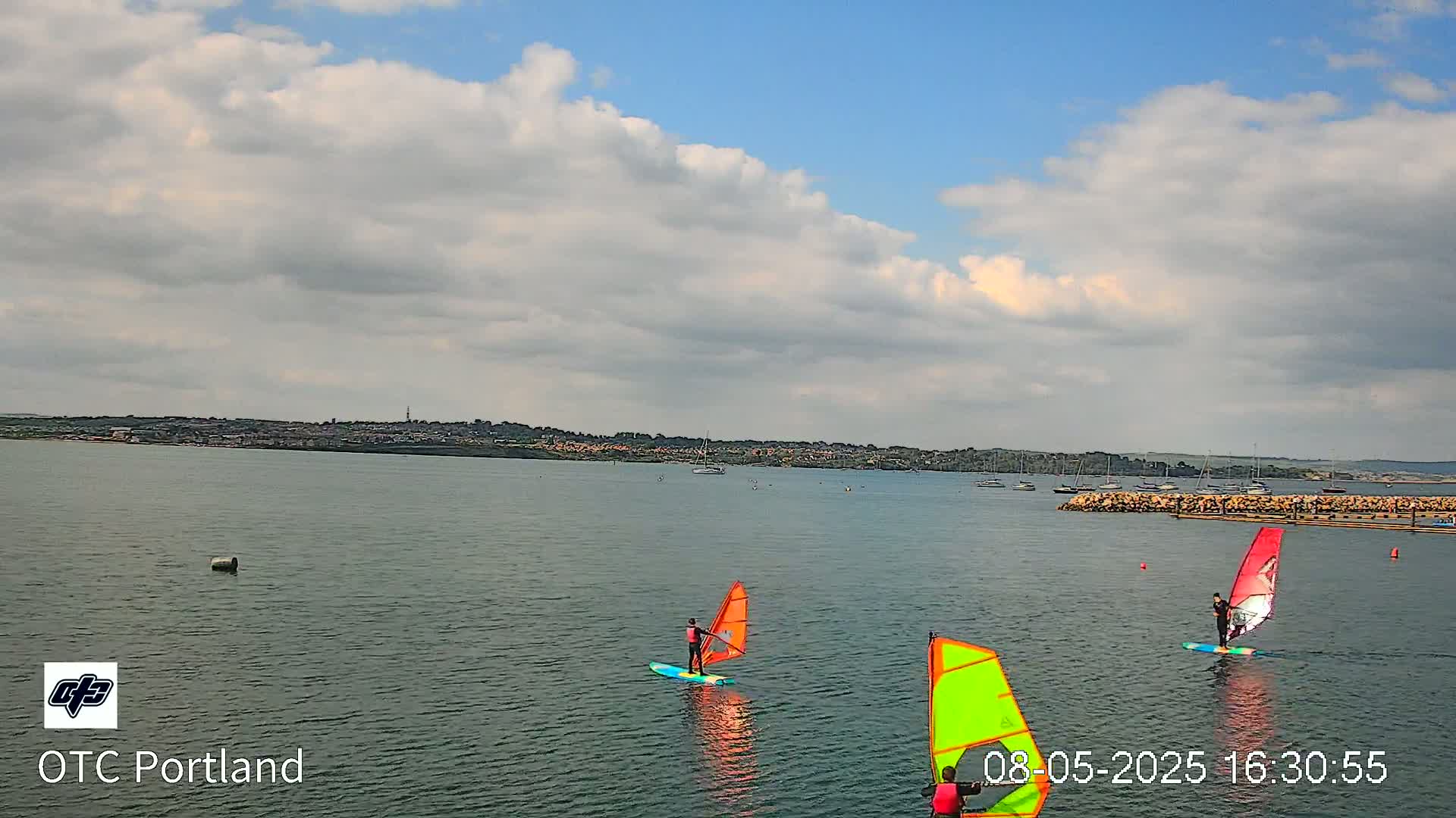 Three windsurfers sail on a calm body of water under a partly cloudy sky.