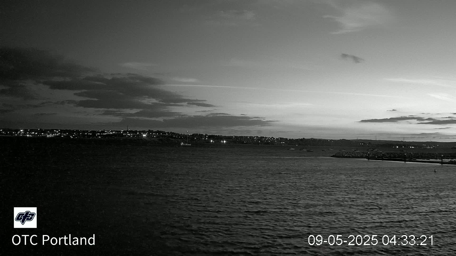 A grayscale image shows a dark, mostly cloudy twilight sky over calm water, with a distant, dimly lit town on the horizon.