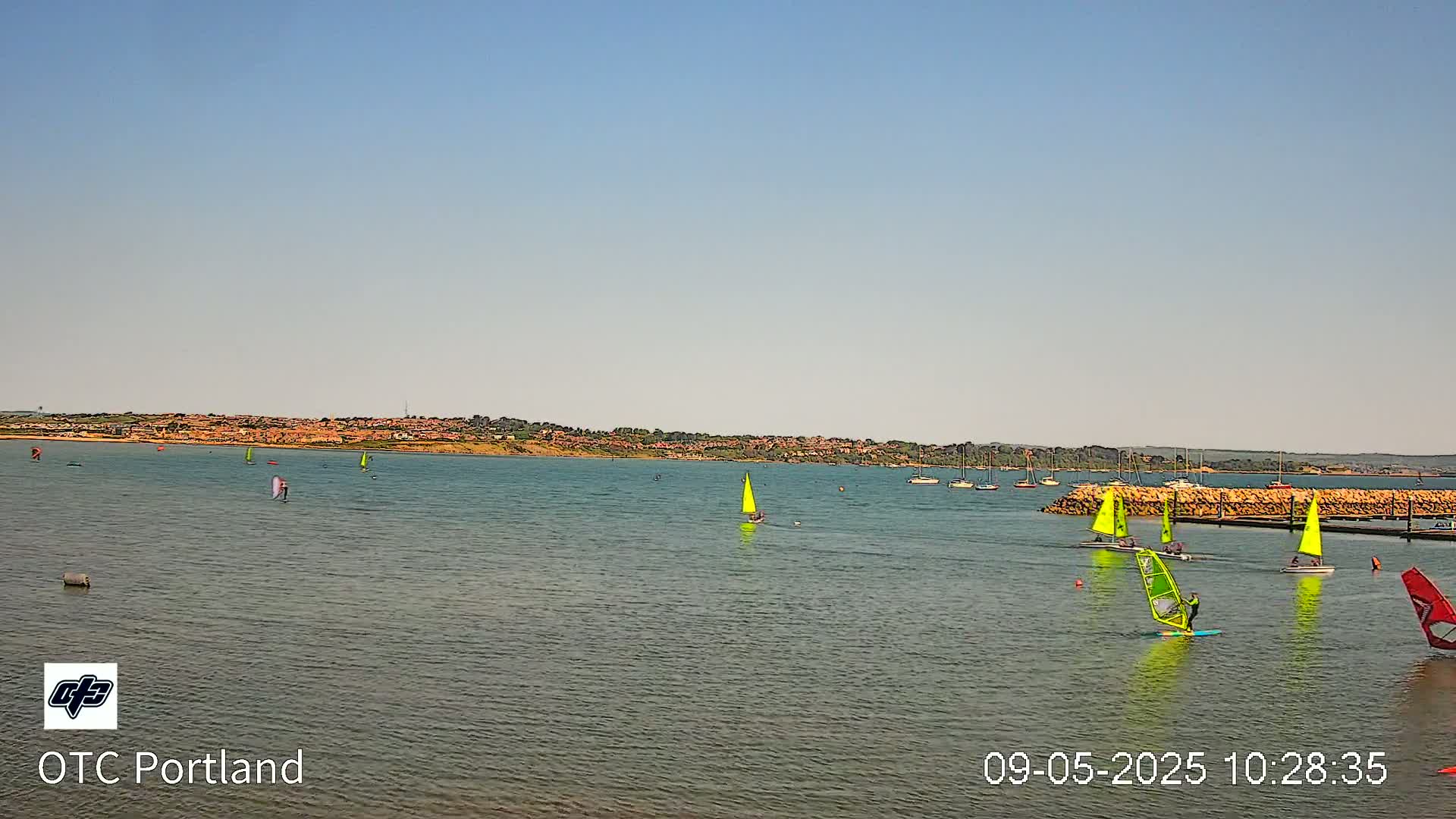 Several sailboats and windsurfers are on a calm body of water with a distant shoreline under a clear sky.
