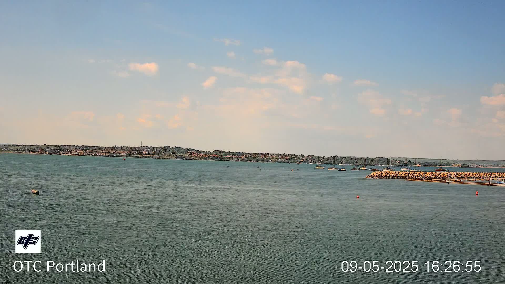 A calm body of water shows a distant shoreline with some buildings and a few sailboats under a partly cloudy sky.