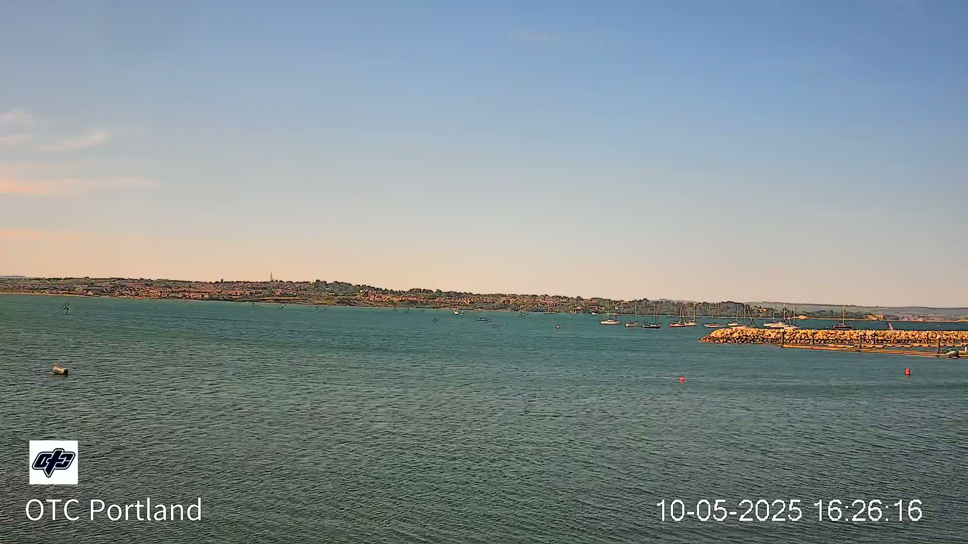 A calm body of water shows several sailboats near a distant shoreline under a clear, light-blue sky.