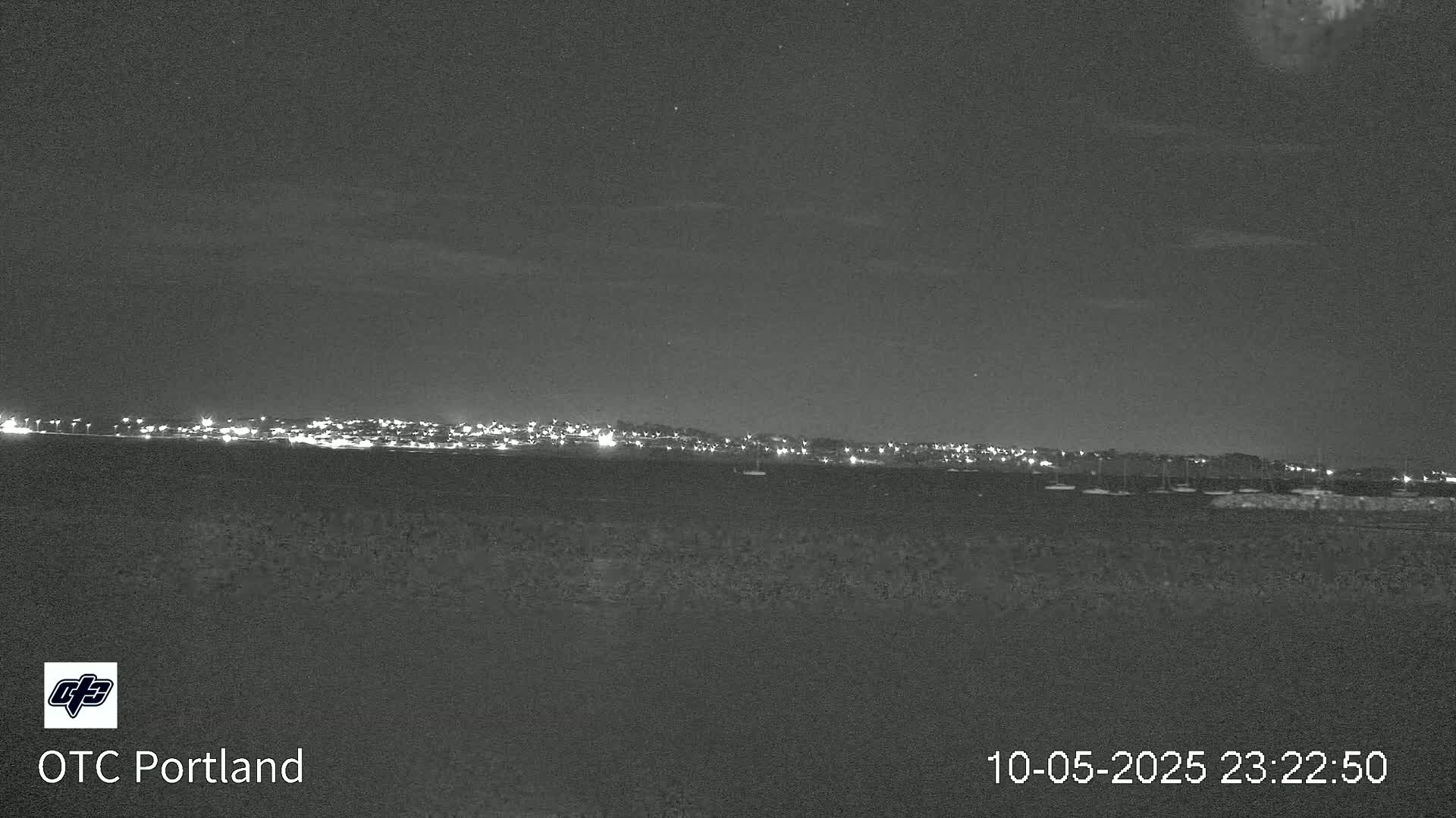 A nighttime long shot shows a distant city skyline illuminated against a dark, clear sky over a body of water.