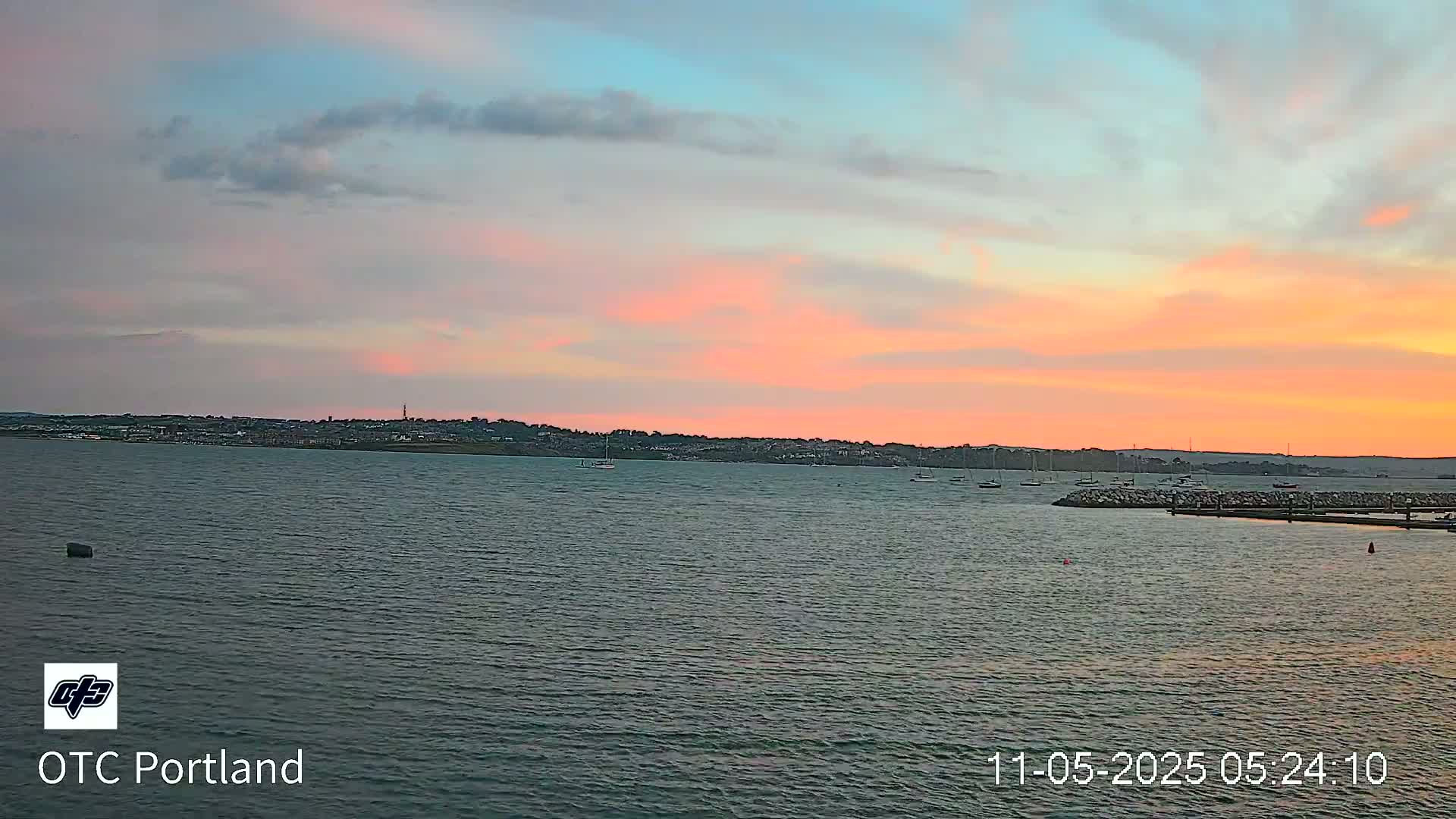 A calm body of water shows a pink and orange sunset over a distant town and a few boats at a marina.
