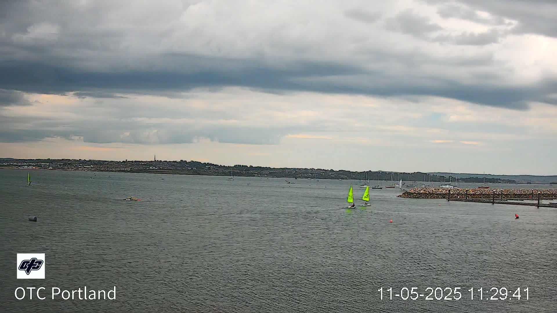 A body of water with several sailboats under a cloudy sky shows a distant shoreline.