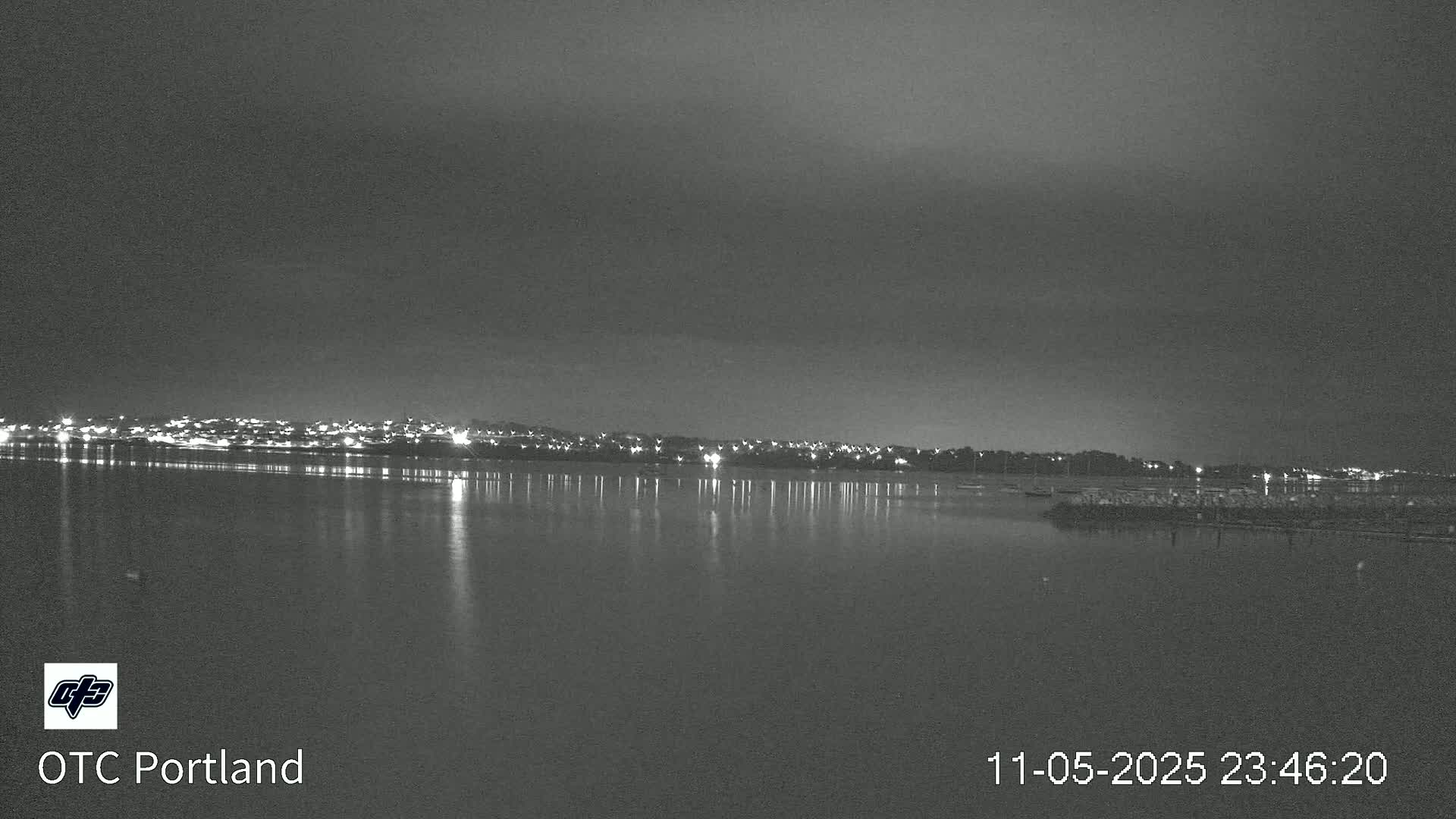 A nighttime view of a calm body of water reflecting the lights of a city across the shore under a dark, cloudy sky.