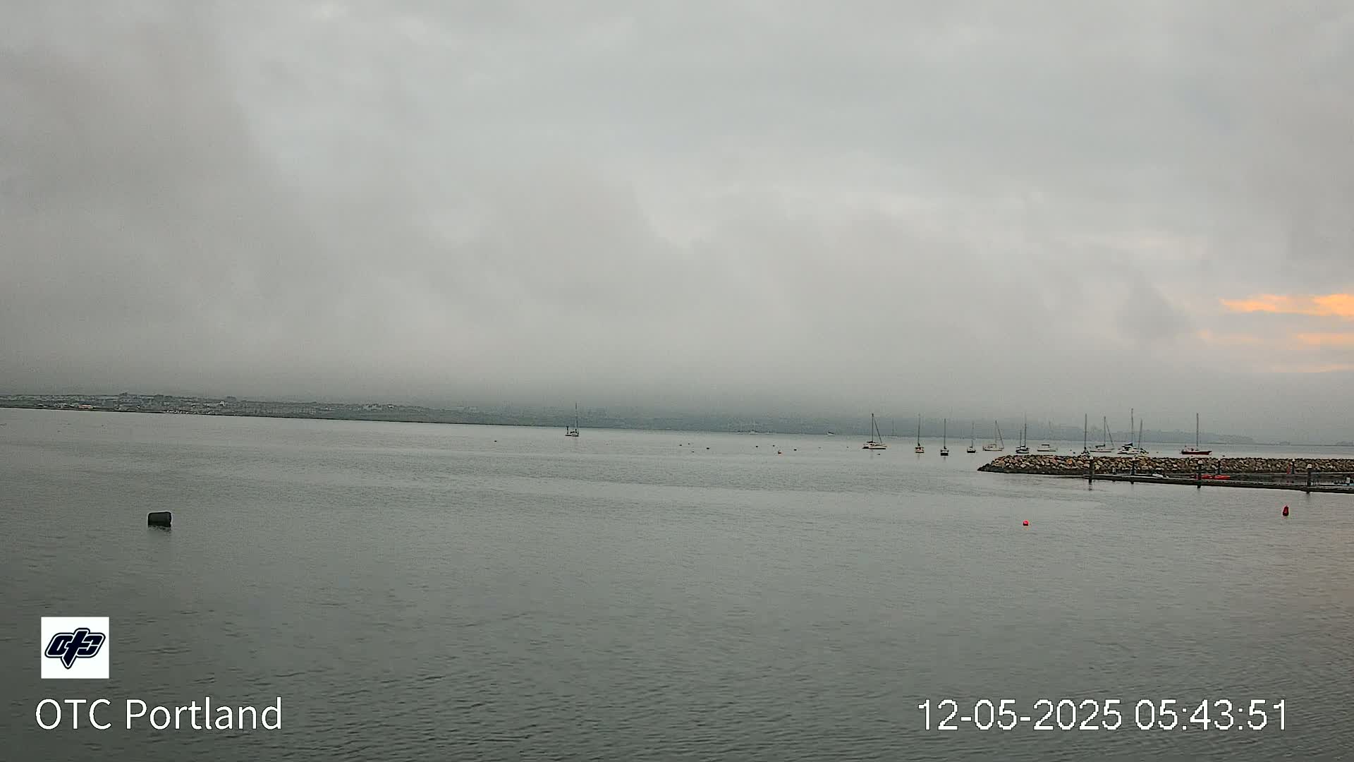 A calm body of water with several boats moored near a rocky shore under a foggy, overcast sky.