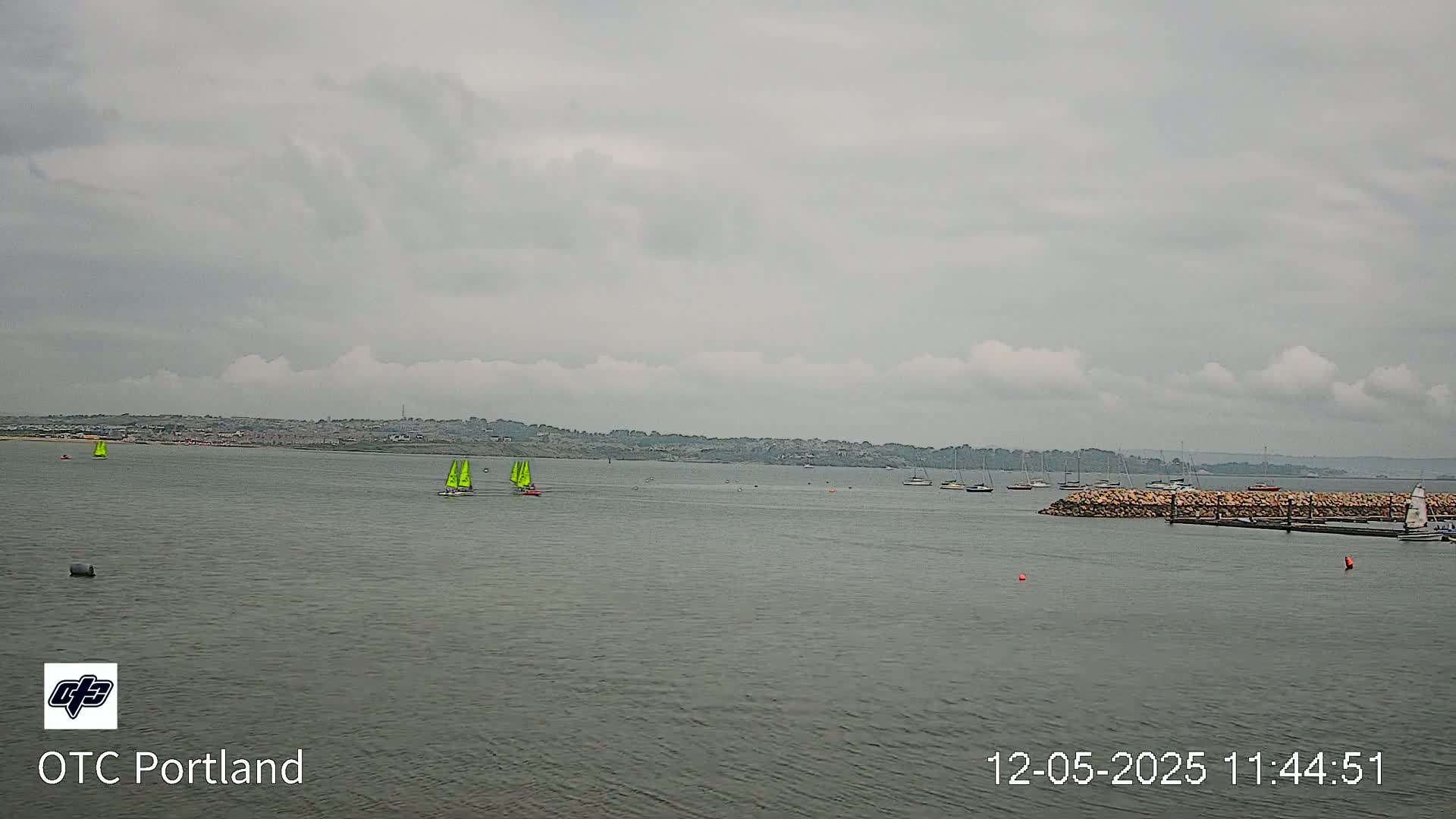 Several sailboats are visible on a calm body of water under an overcast sky, with a distant shoreline in the background.