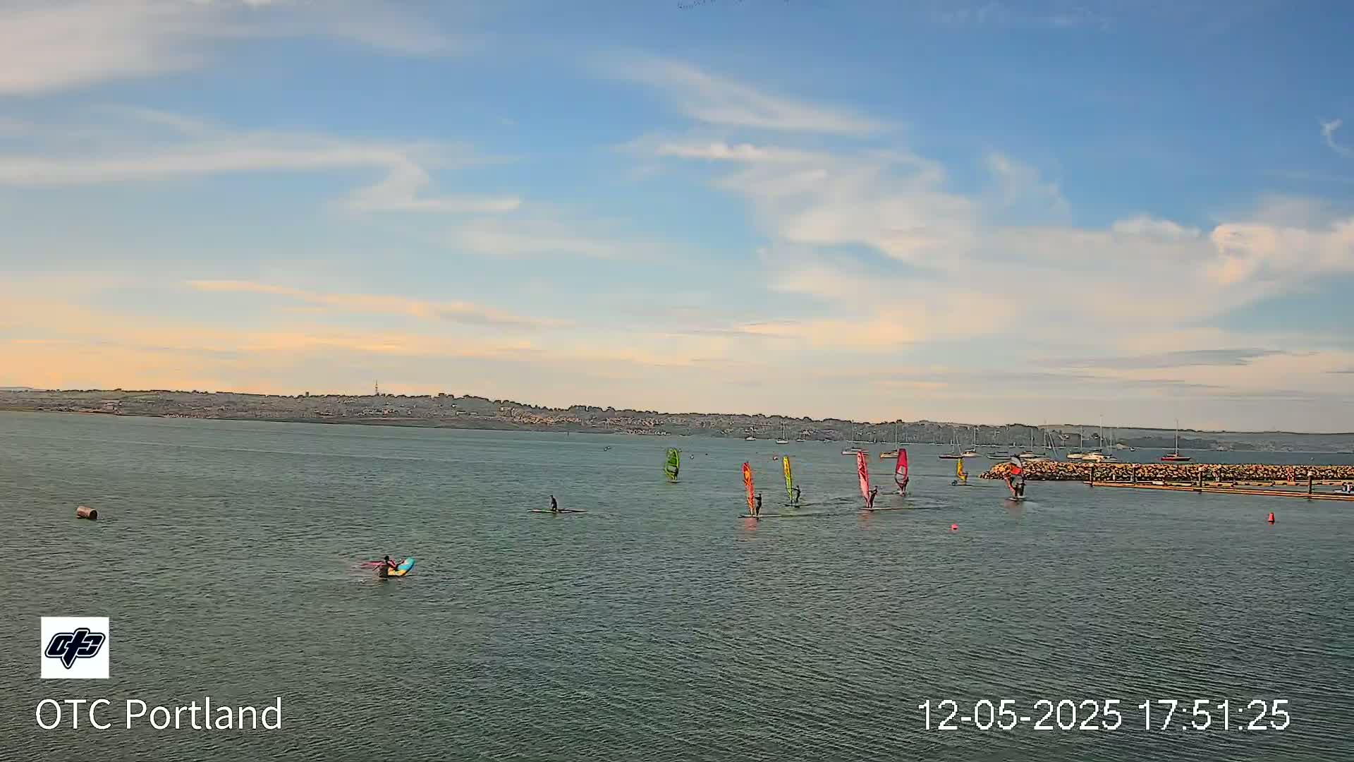 Several windsurfers sail on a calm body of water under a partly cloudy sky.