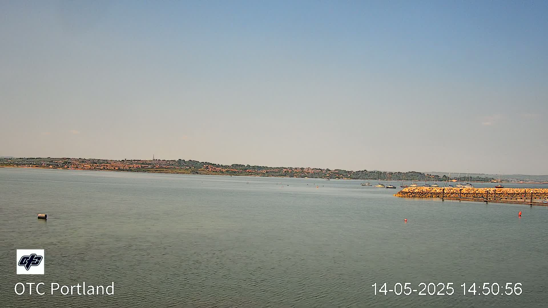 A calm body of water shows a distant shoreline with buildings and trees under a clear sky.