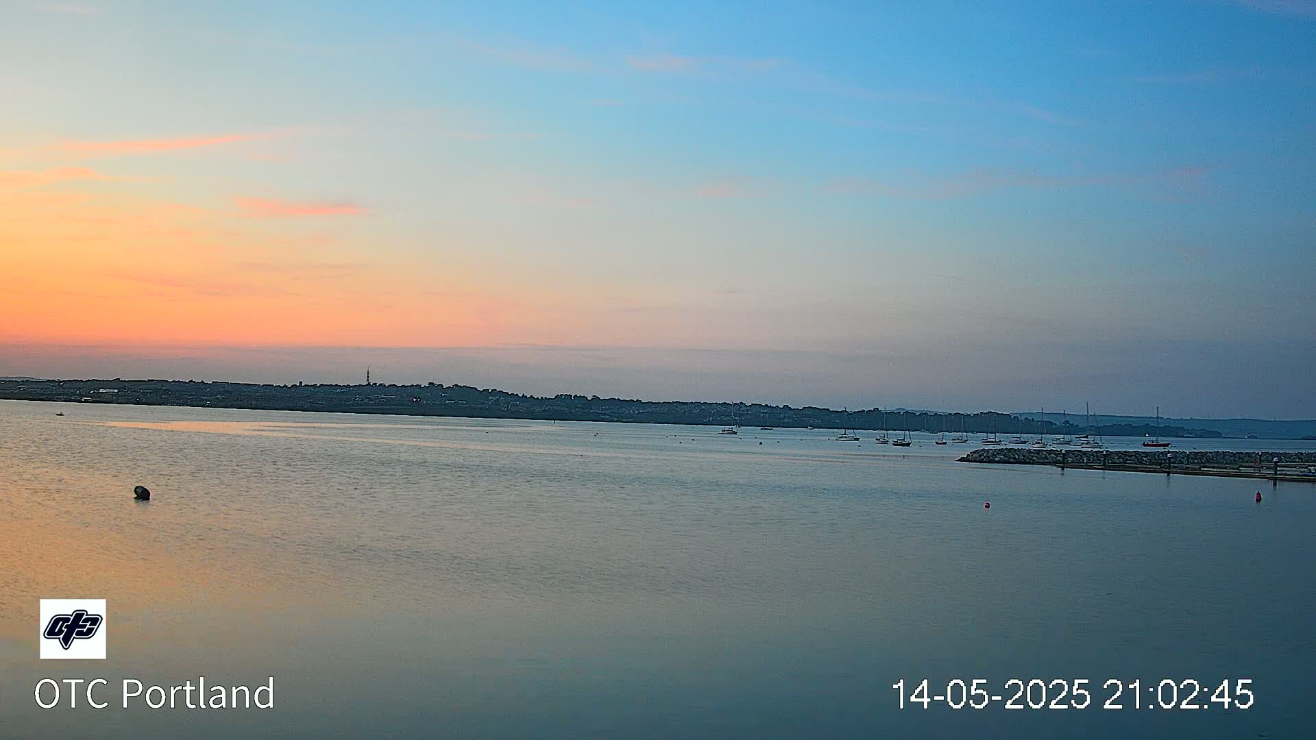 A calm body of water with several boats at anchor is seen at sunset under a pastel sky.