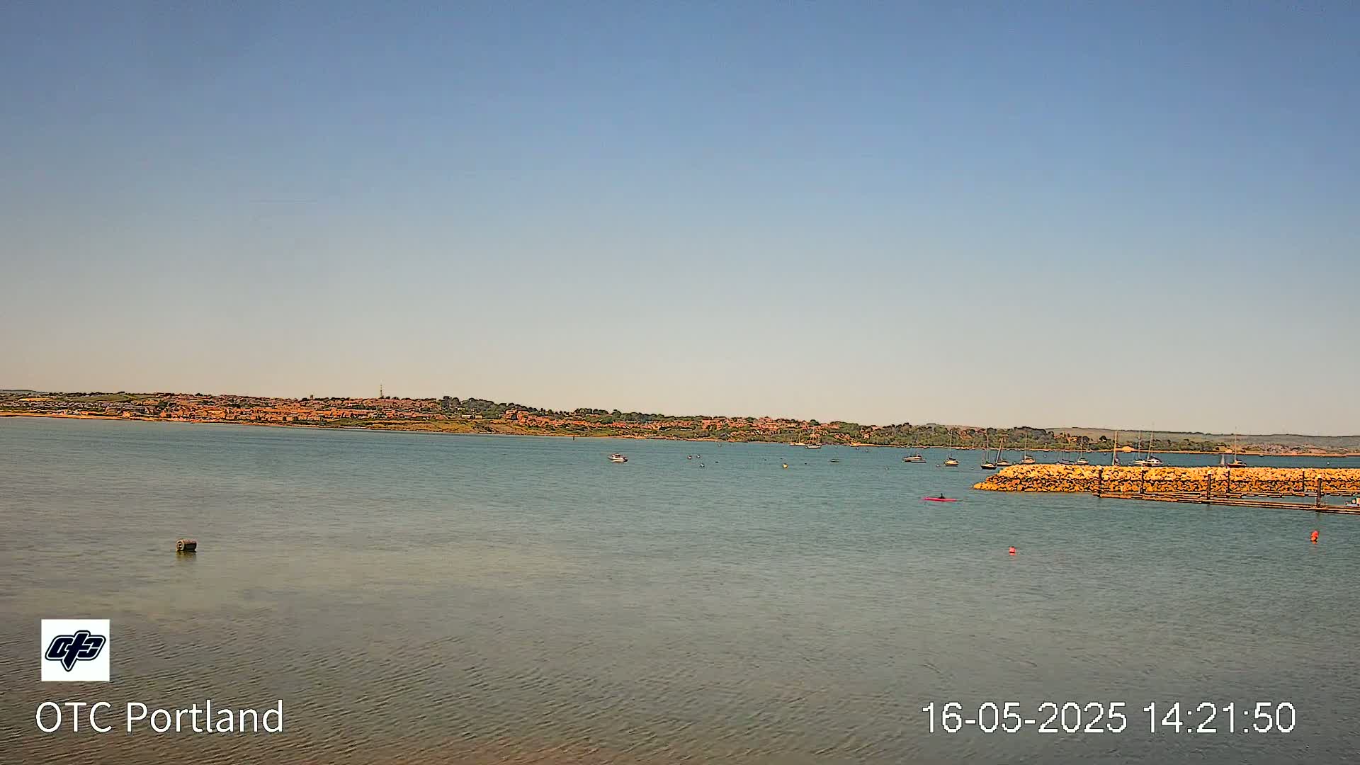 A calm body of water shows a distant shoreline with buildings under a clear, light-blue sky, and a few boats and a kayak are visible on the water.