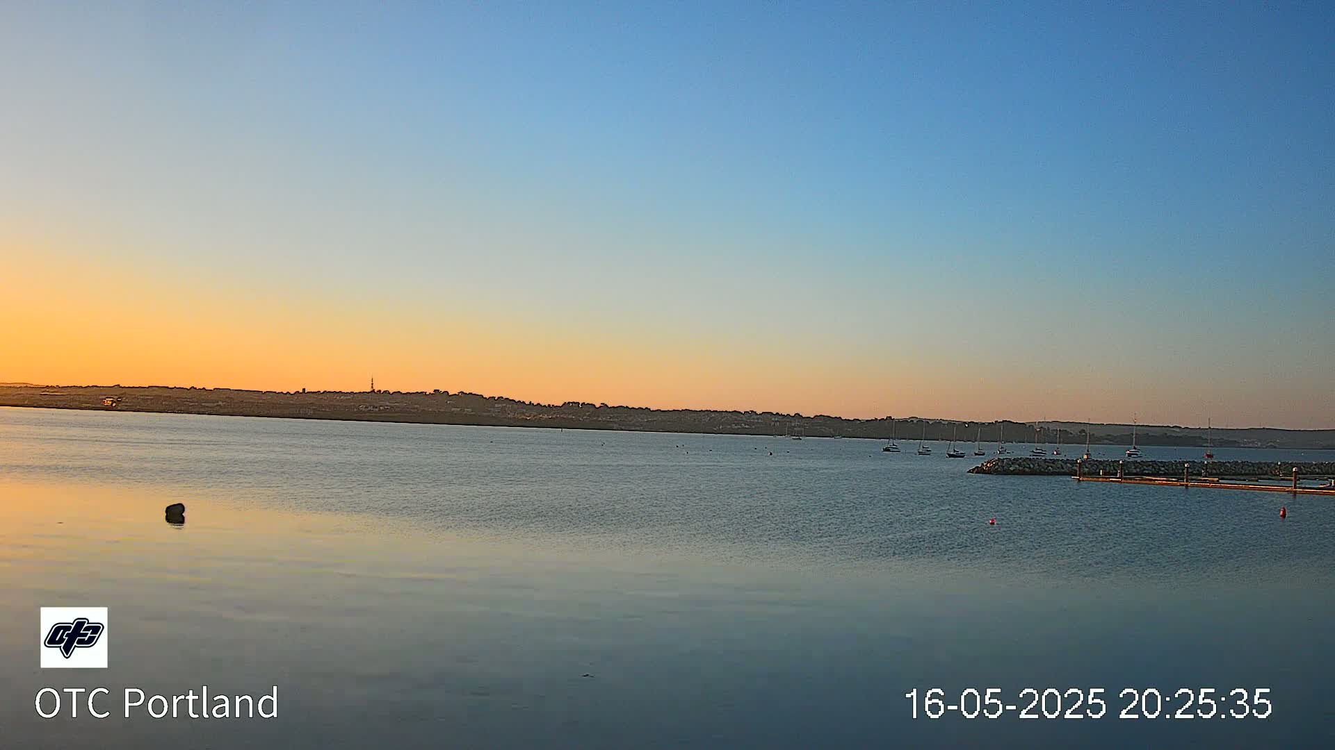 A calm body of water at sunset shows a few sailboats near a shoreline with a distant landmass under a clear, light-blue sky.