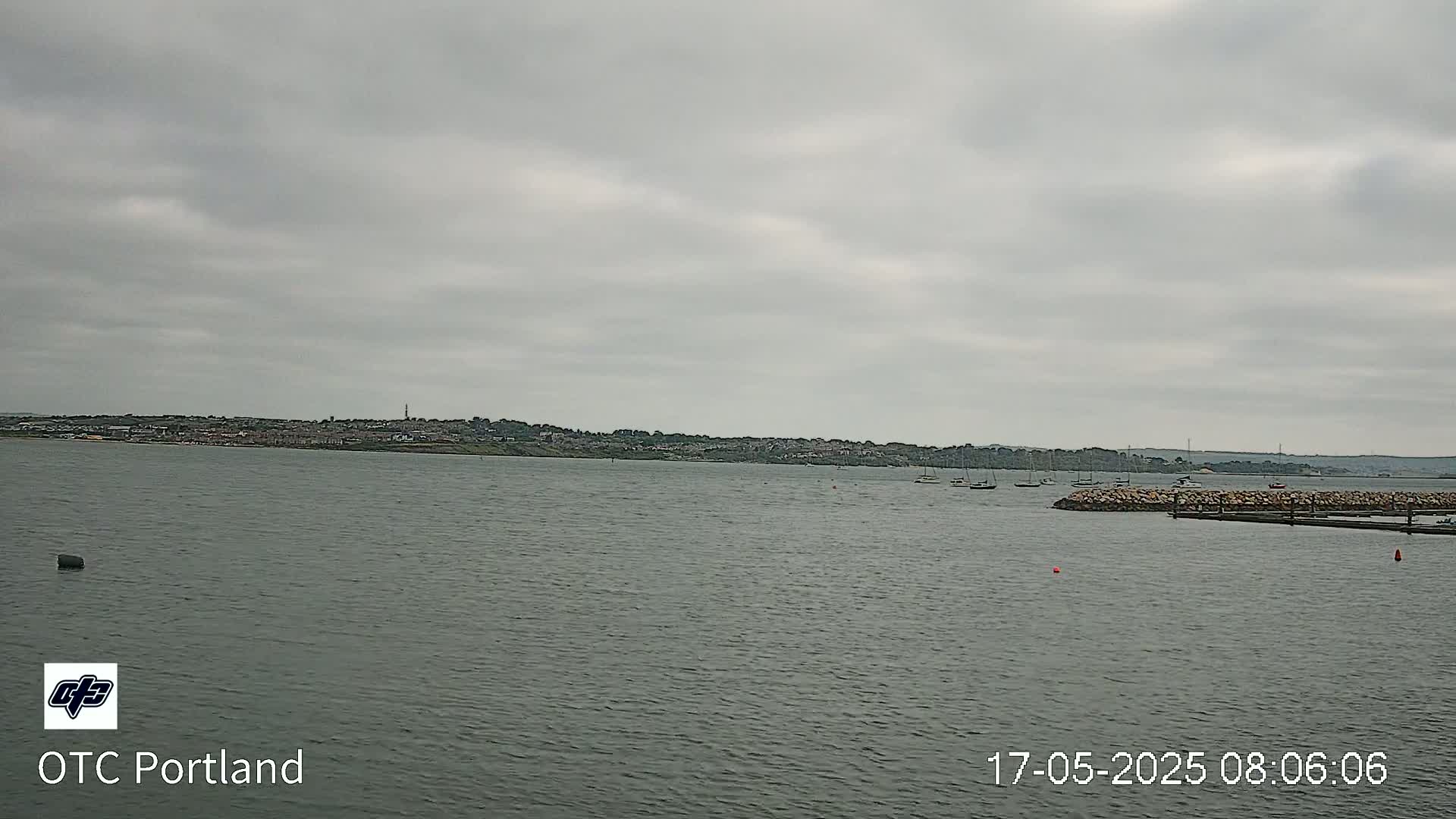 A calm body of water shows a distant town under an overcast sky, with several boats moored near a rock jetty.