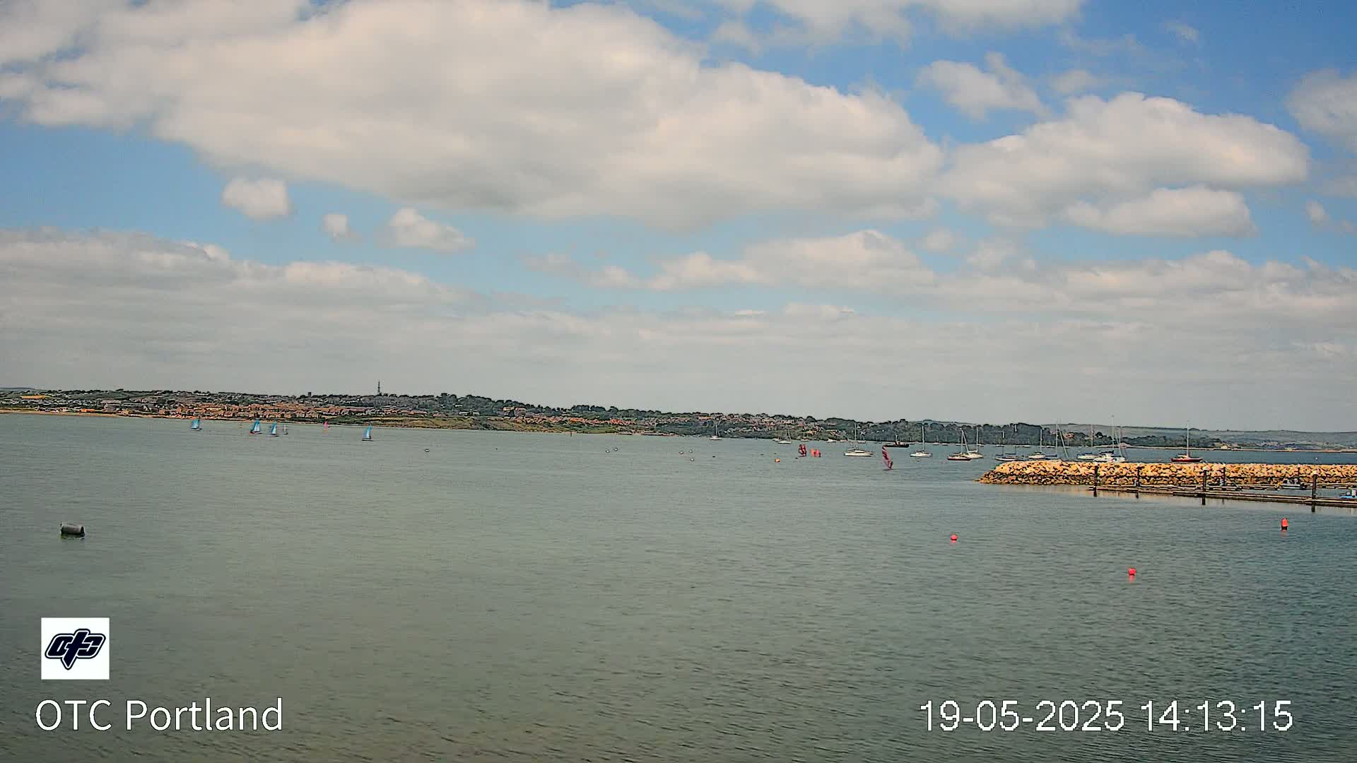 A partly cloudy day shows sailboats on a calm body of water in front of a distant shoreline.