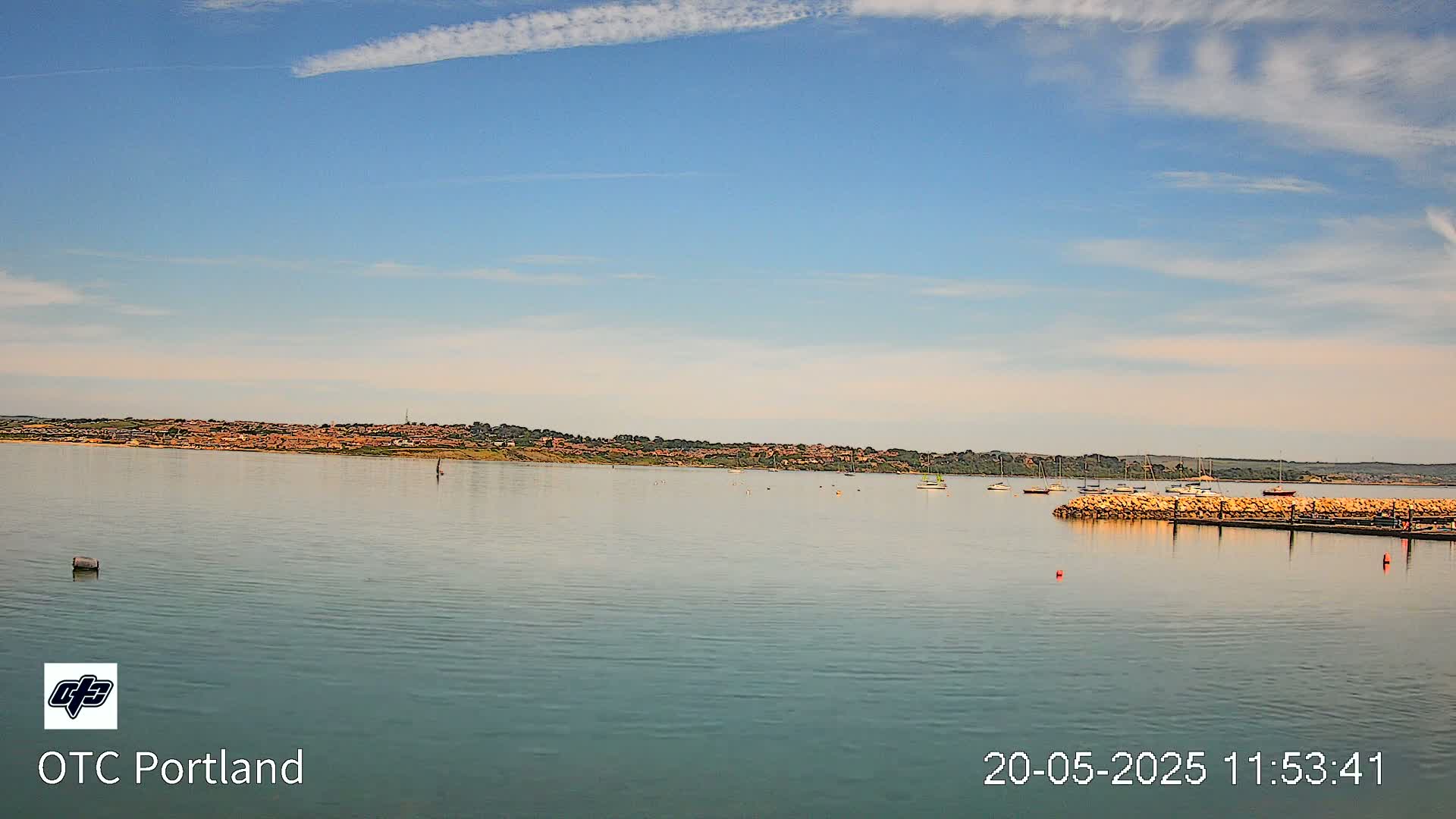 A calm body of water shows a distant shoreline with buildings under a mostly clear, light blue sky.