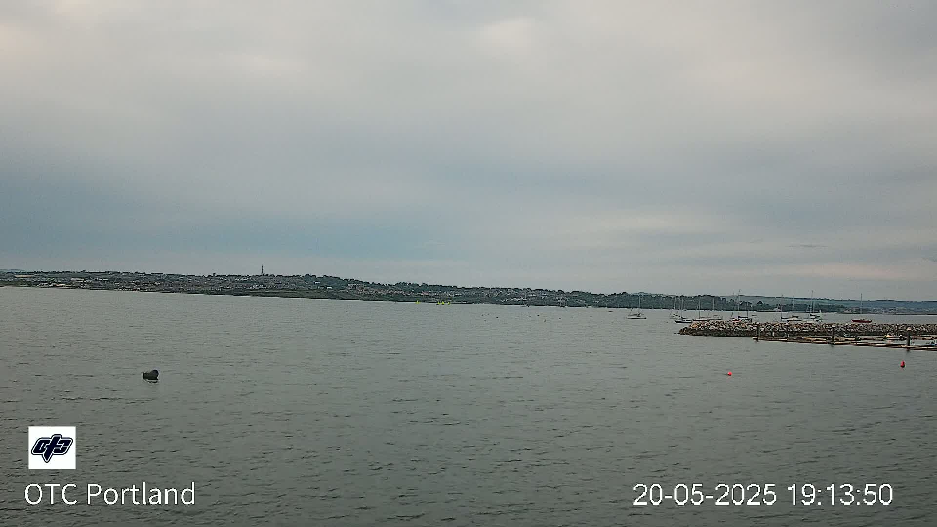 A calm body of water stretches out to a distant shoreline under an overcast sky, with several boats moored near a rocky breakwater.
