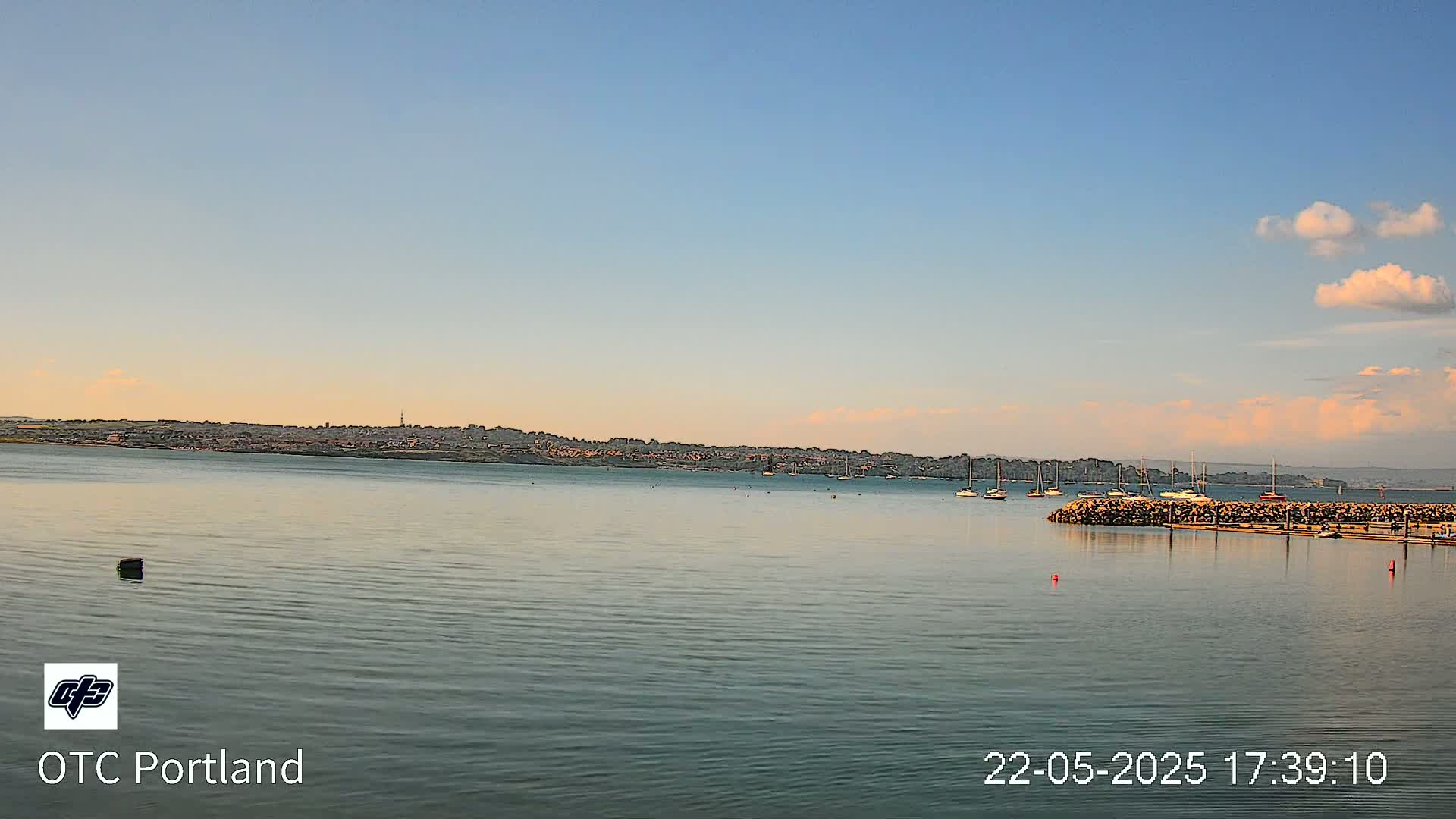 A calm body of water shows a distant town under a mostly clear sky with a few puffy clouds.