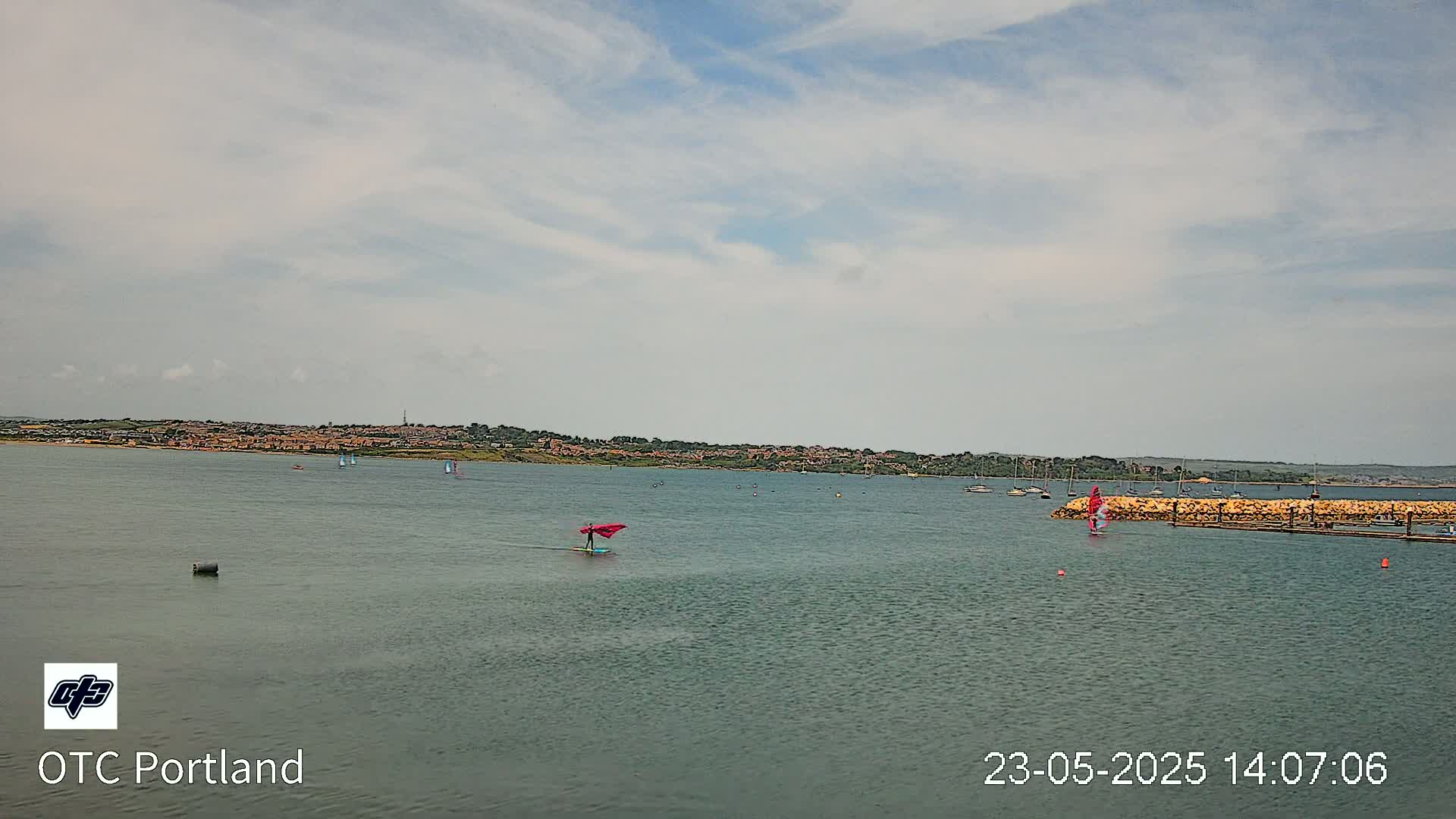 A calm body of water with several sailboats and windsurfers, under a partly cloudy sky, shows a distant shoreline with buildings.