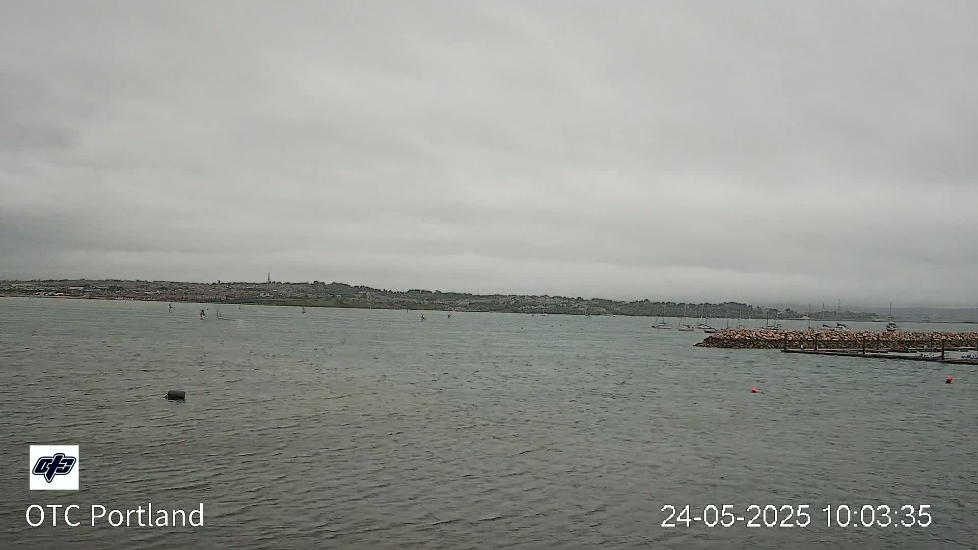 A calm body of water with several sailboats and windsurfers is visible under an overcast sky, with a distant shoreline in view.