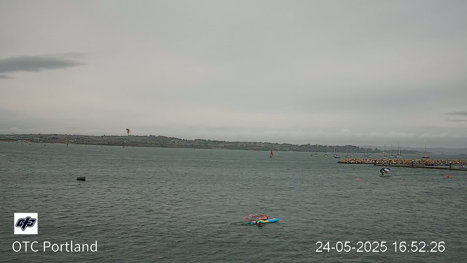 A wide shot of a body of water on an overcast day shows several sailboats and windsurfers, with a distant shoreline visible in the background.