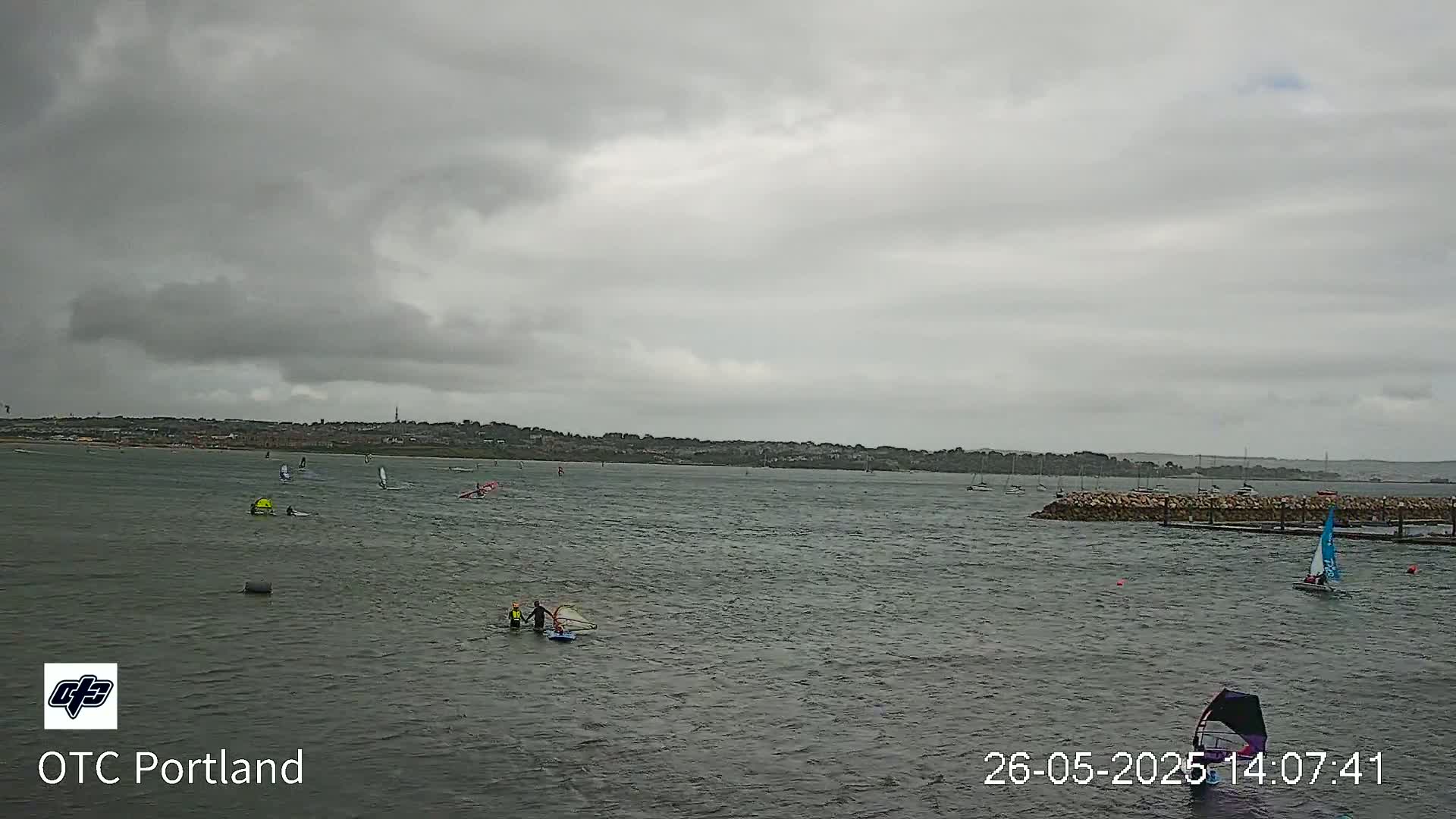 Several windsurfers are on the water under an overcast sky, with a distant shoreline visible.
