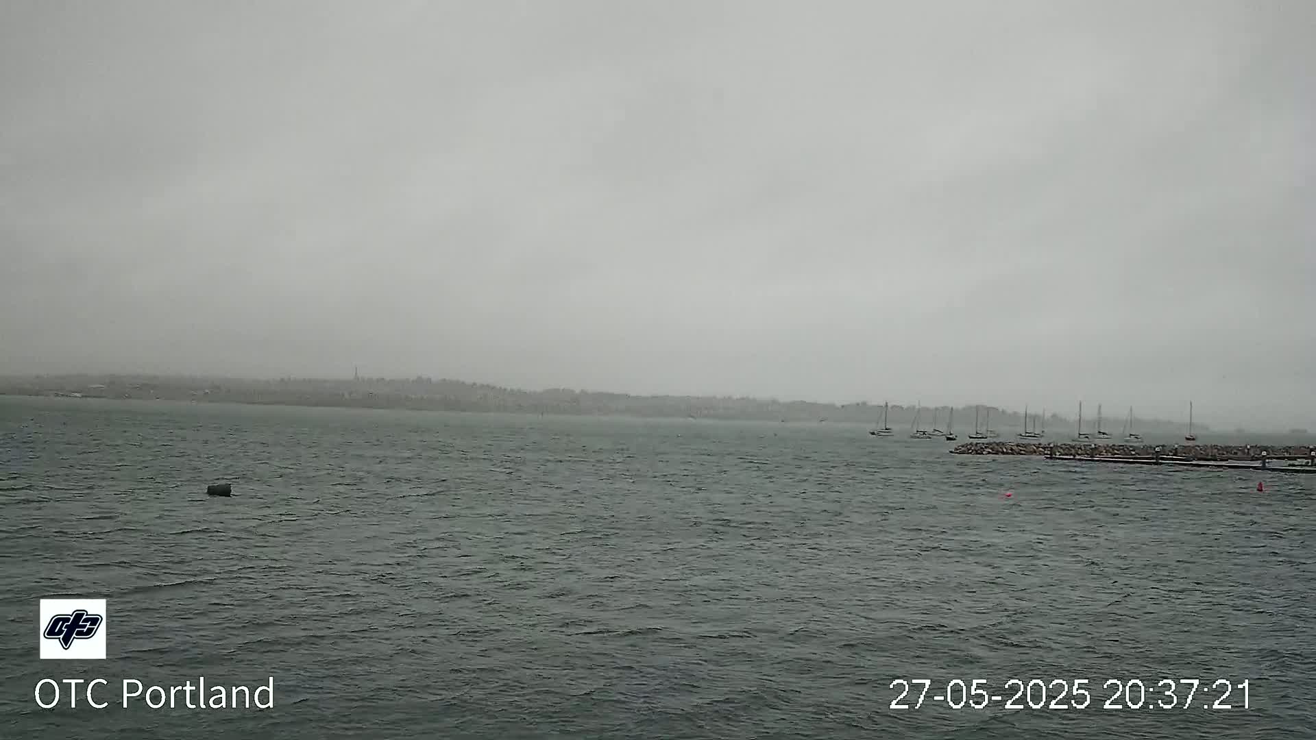 A gray, overcast day shows a choppy body of water with several sailboats moored near a rocky shoreline and a distant landmass under a heavy, grey sky.