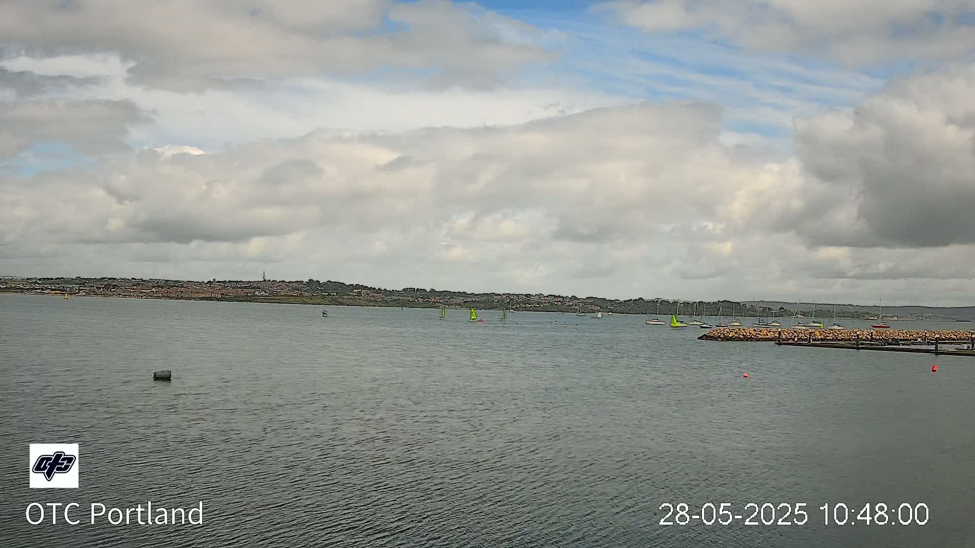 A body of water with several sailboats and a distant shoreline under a mostly cloudy sky.