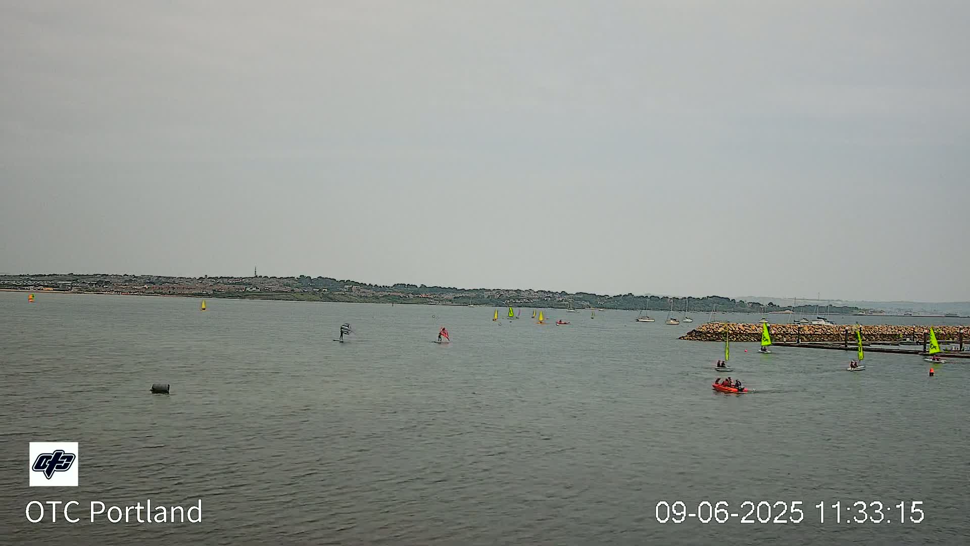 Several sailboats and windsurfers are on a calm, gray day on a body of water with a distant shoreline.
