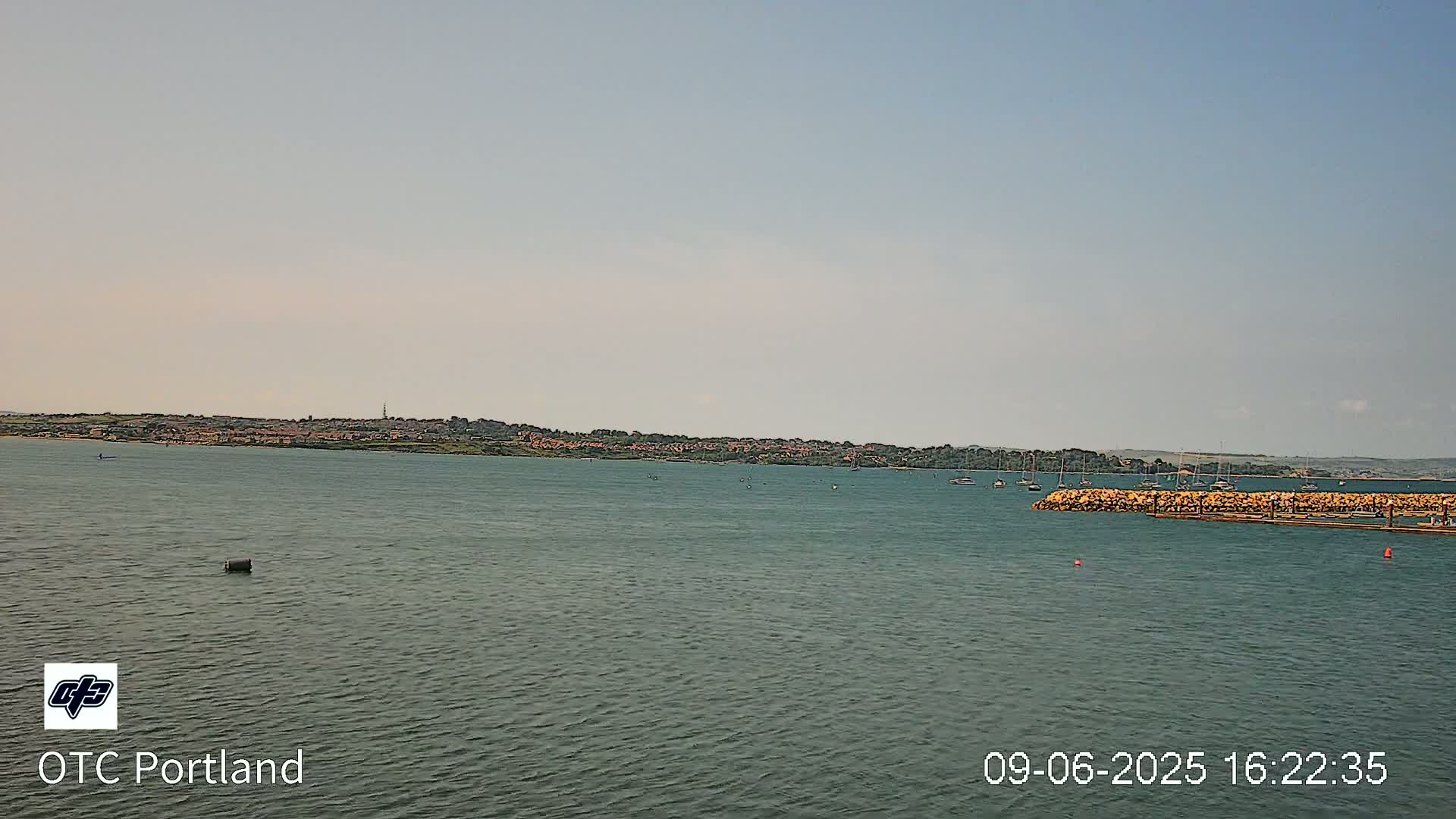 A calm body of water shows a distant shoreline with a few boats and a breakwater under a clear sky.