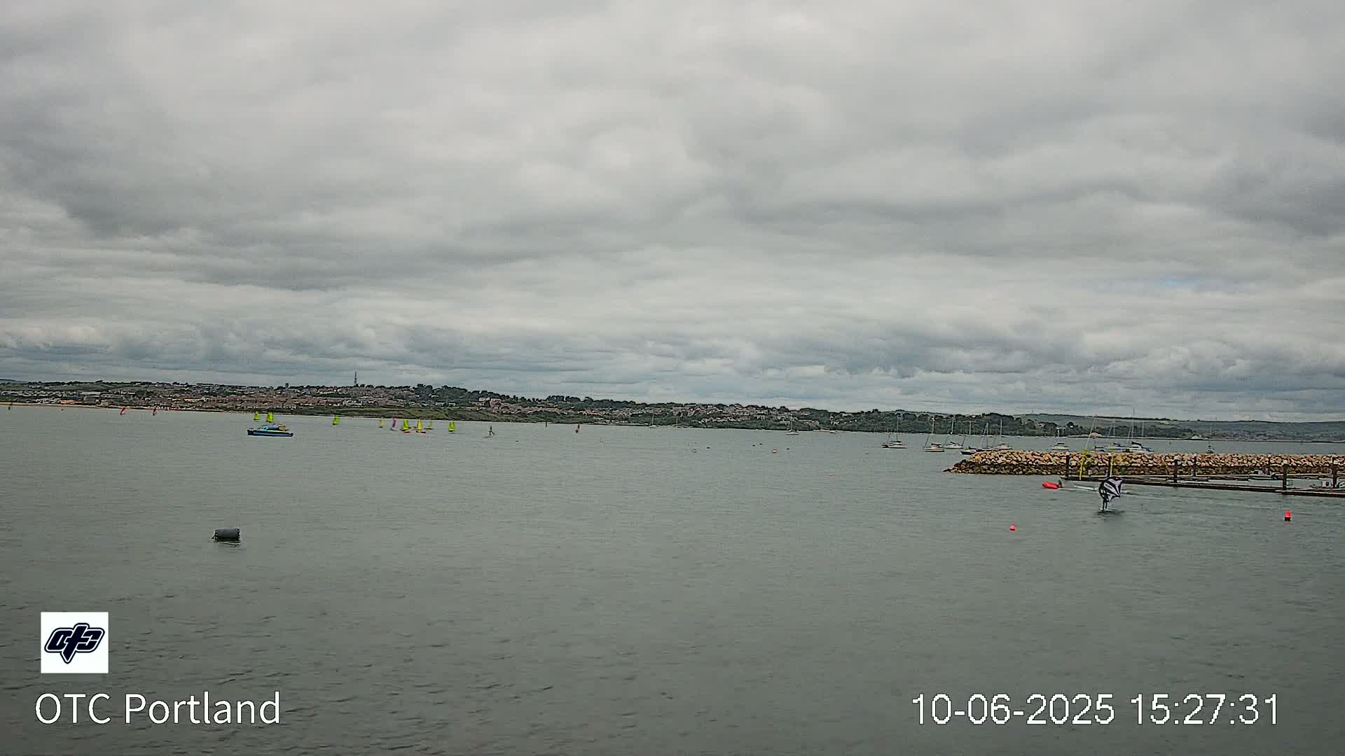 A calm body of water with several sailboats and a person windsurfing under an overcast sky.