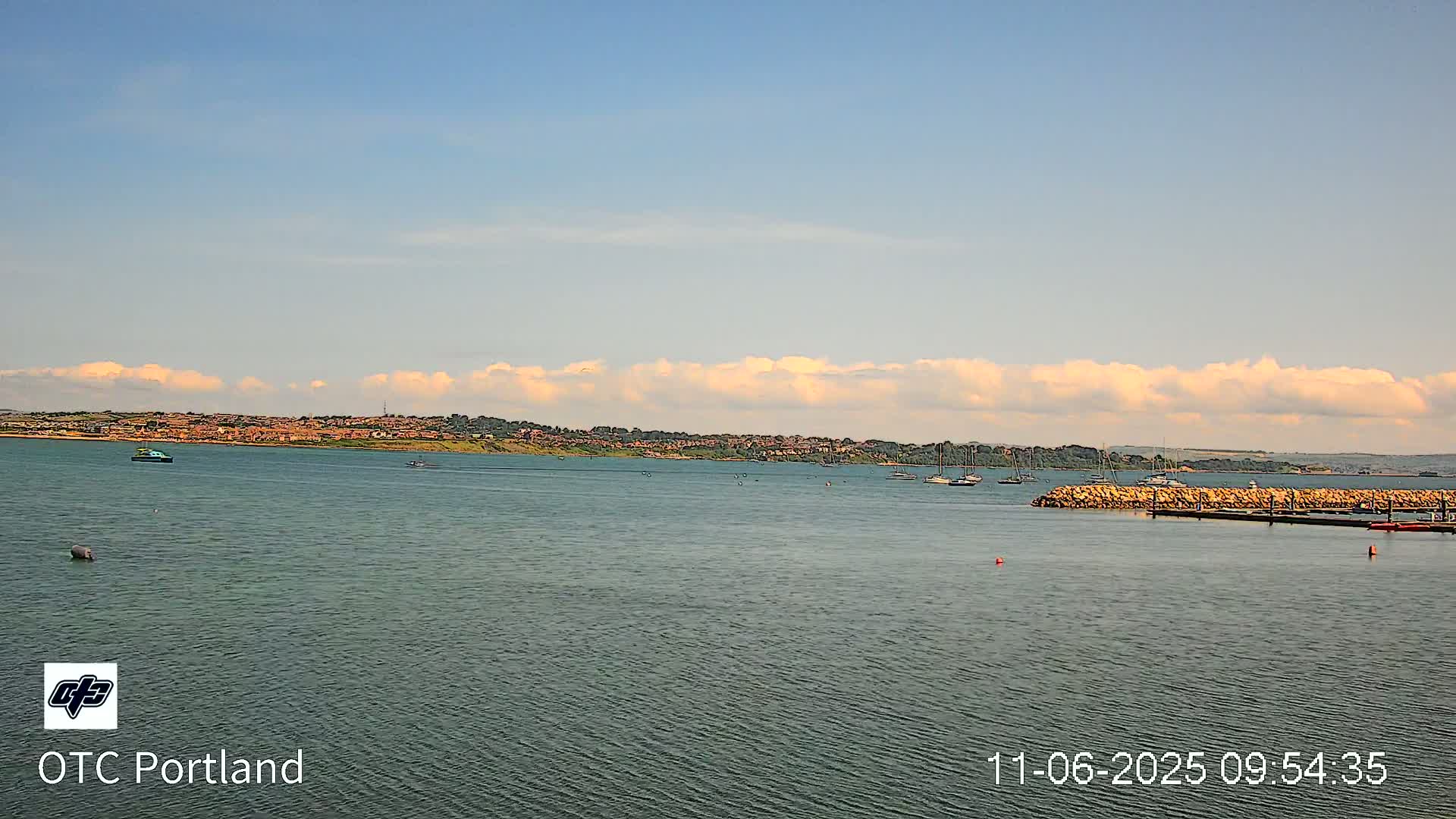 A calm body of water with a few boats, a distant shoreline with buildings, and a mostly clear sky with some fluffy clouds.