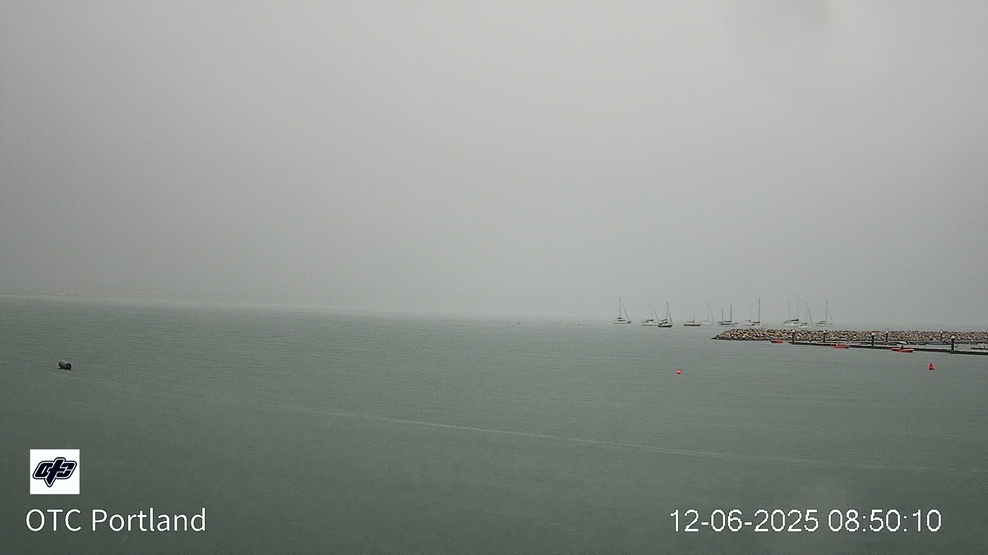A hazy, gray day shows a calm body of water with several sailboats moored near a stone jetty.