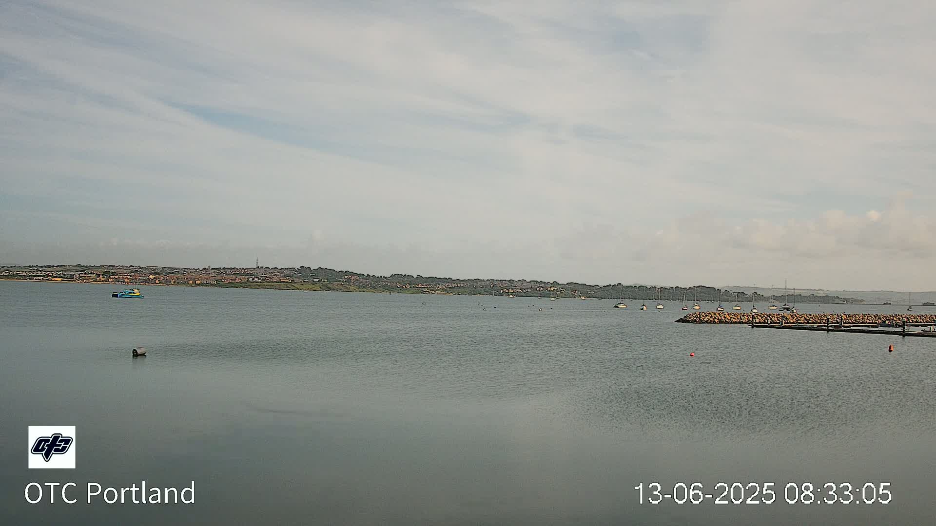 A calm body of water shows a distant shoreline with several small boats and a rock jetty under an overcast sky.