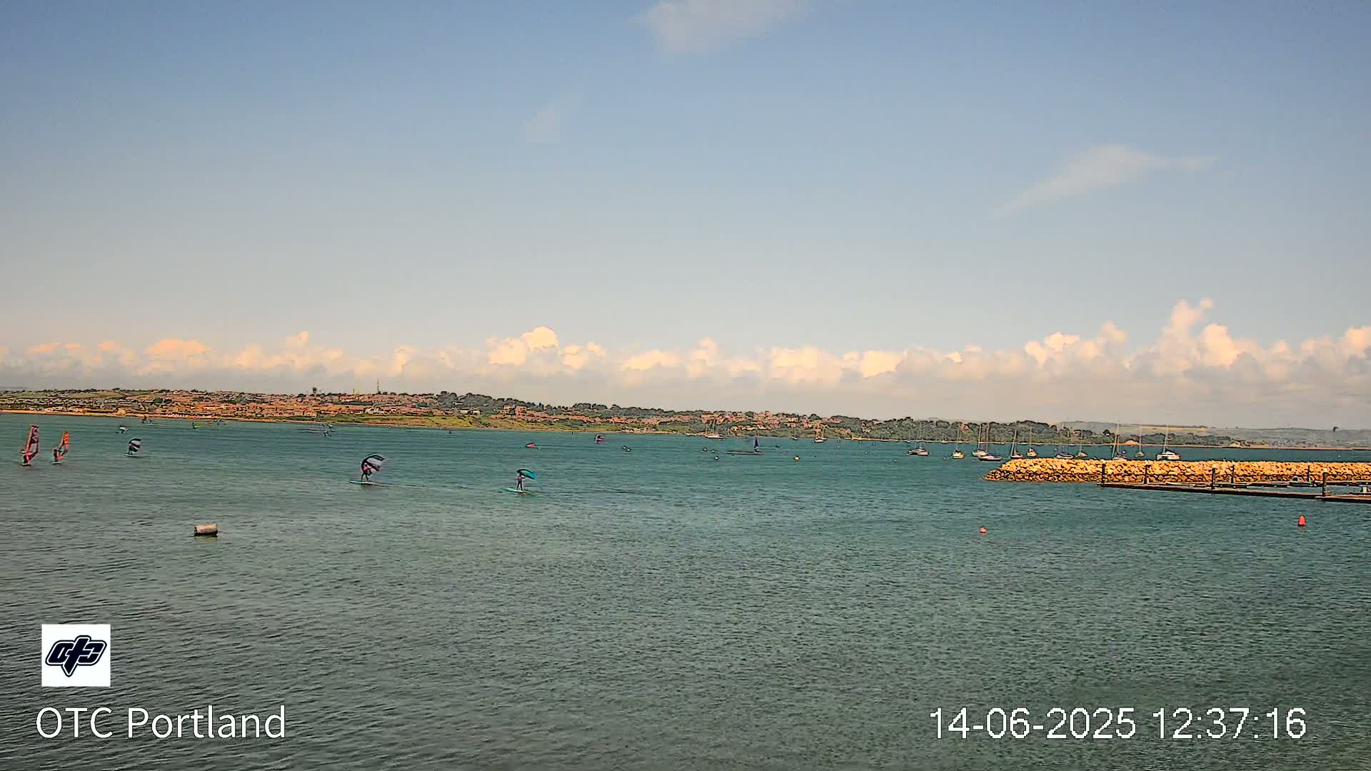 Several windsurfers and sailboats are visible on a calm body of water under a partly cloudy sky, with a distant shoreline in view.