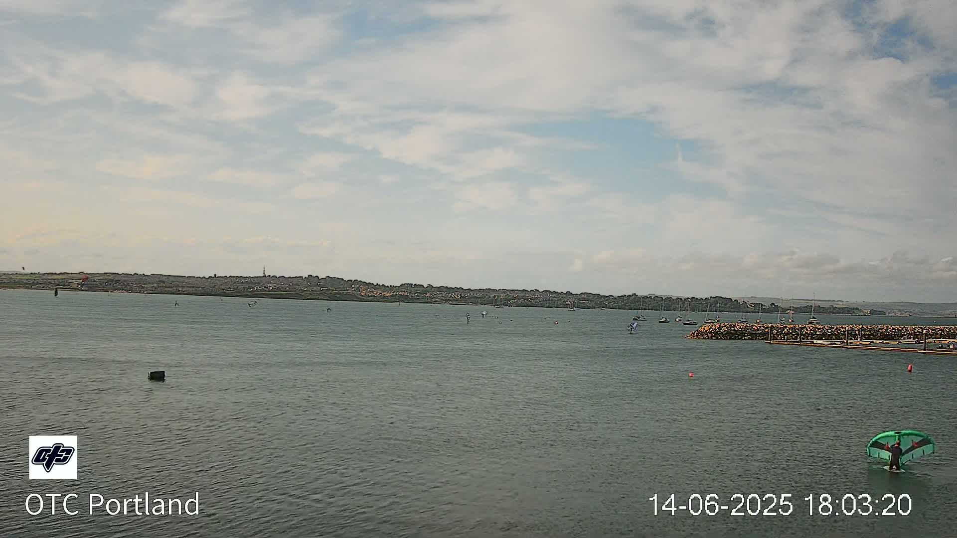 A calm body of water with a few sailboats and a person kitesurfing under a partly cloudy sky.