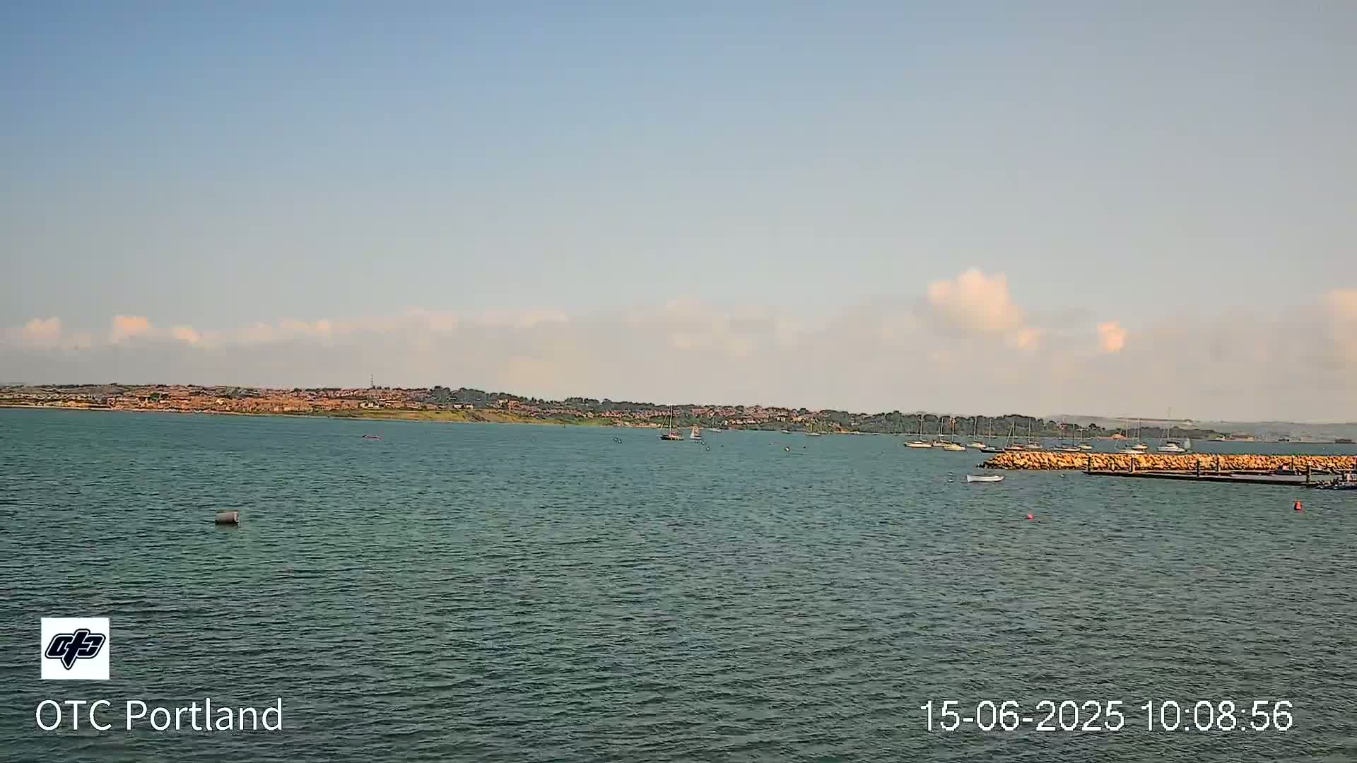 A calm body of water shows a distant shoreline with buildings under a mostly clear sky with a few scattered clouds.