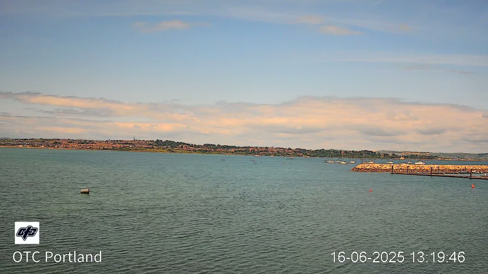 A calm body of water with several boats near a small stone jetty, under a partly cloudy sky, shows a distant shoreline of buildings and trees.
