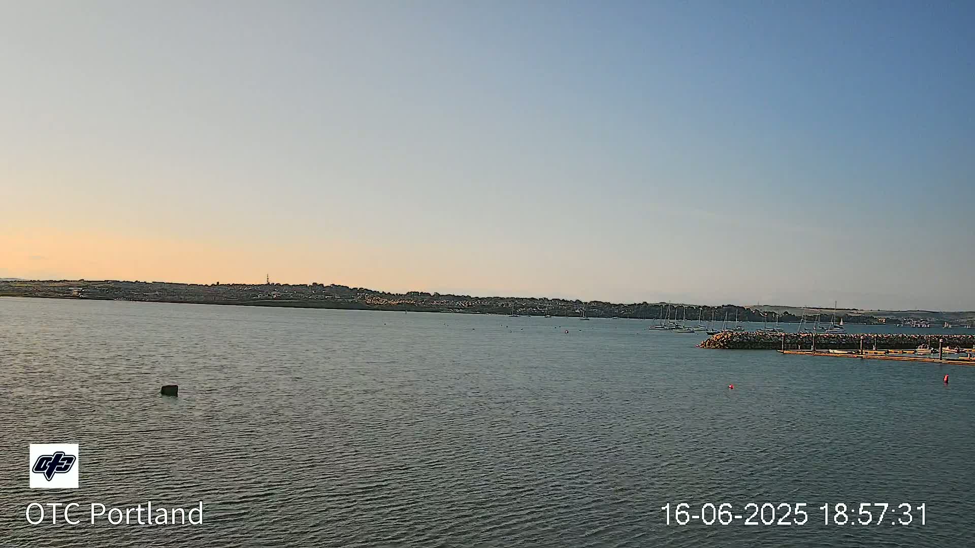 A calm body of water shows a distant shoreline with several sailboats moored near a rock jetty under a clear, twilight sky.