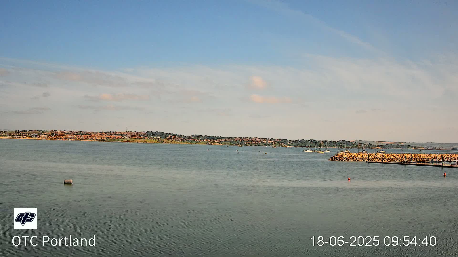 A calm body of water shows a distant shoreline with buildings under a mostly clear sky with a few clouds.
