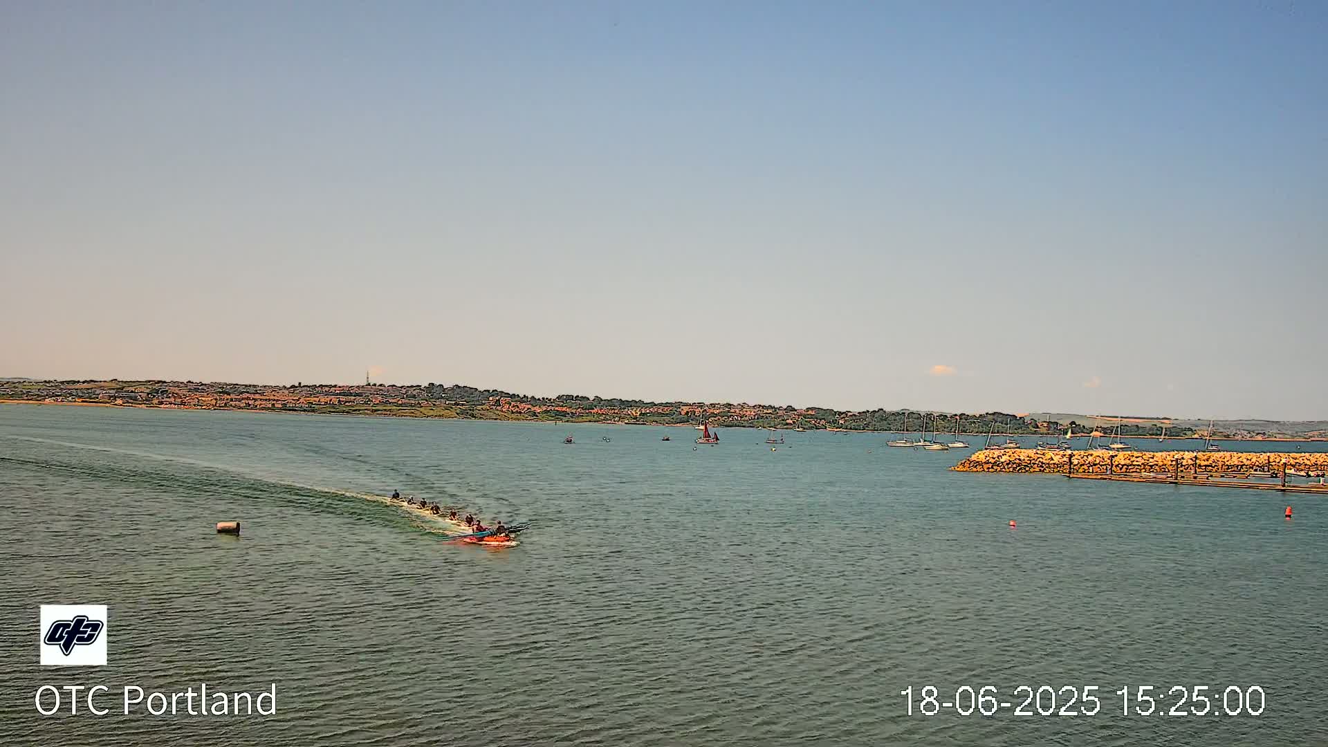 A long, slender boat carrying many rowers travels across calm, green water toward a distant shoreline under a clear, light-blue sky.
