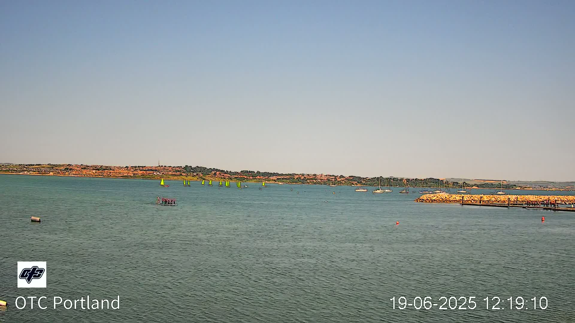 Under a clear sky, a body of water shows several sailboats and a group of people in a small boat, with a distant shoreline visible in the background.
