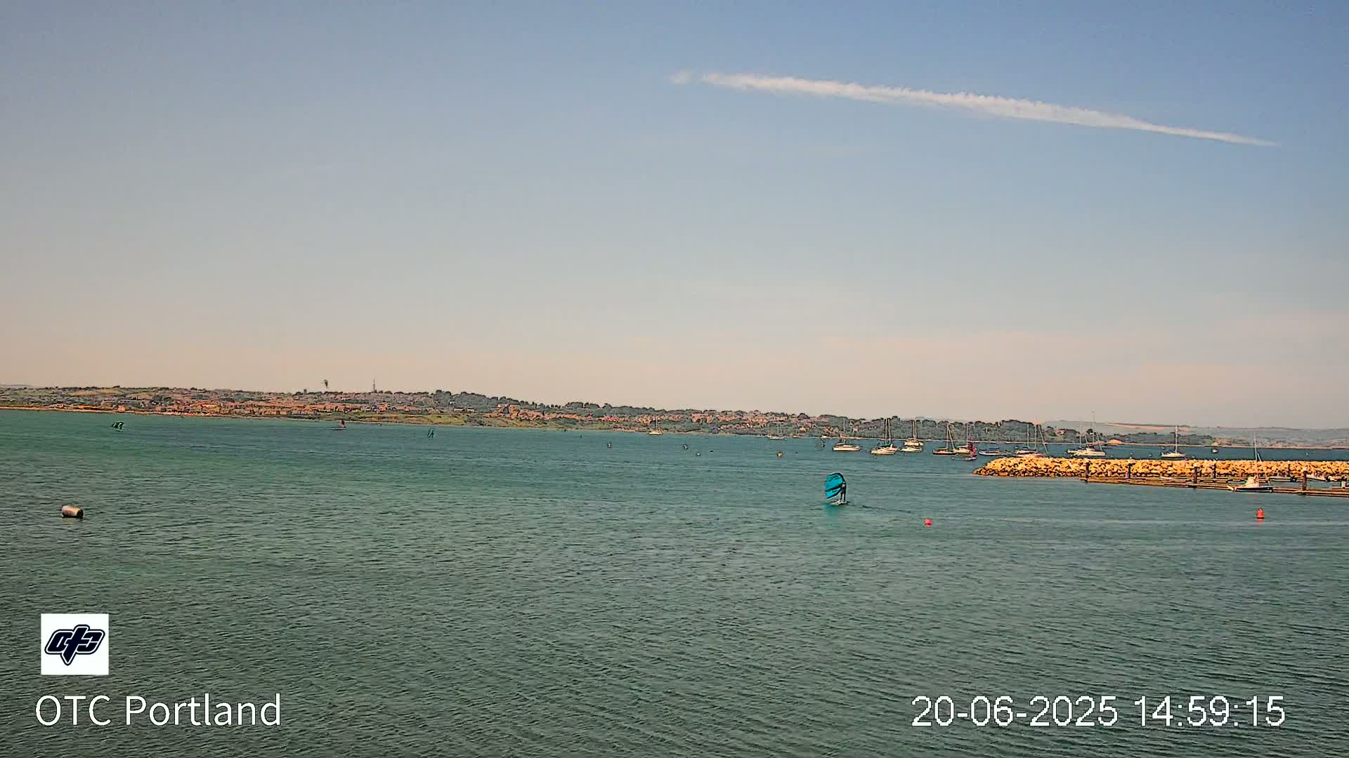 A calm body of water shows several sailboats and windsurfers under a mostly clear sky.