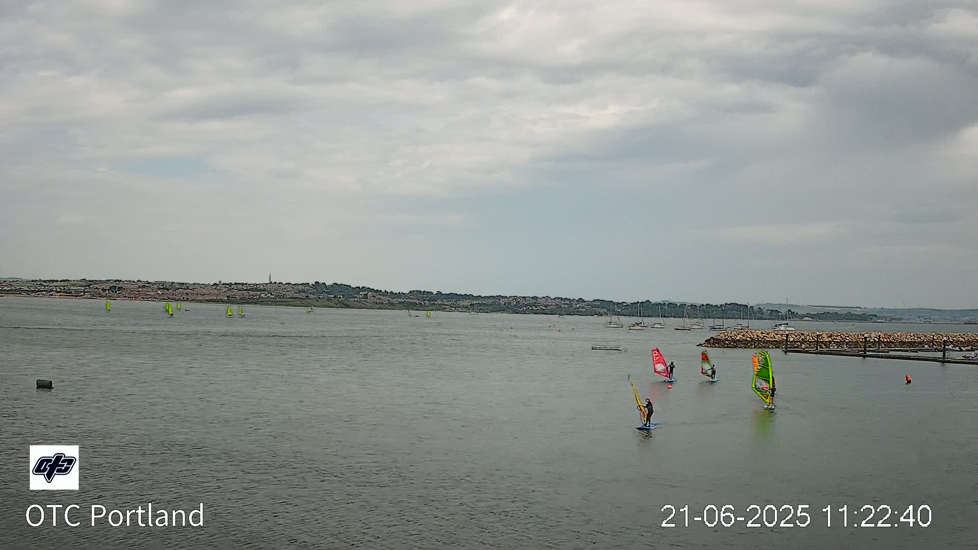 Several windsurfers sail on a calm, gray-cloudy day across a body of water in front of a distant shoreline.