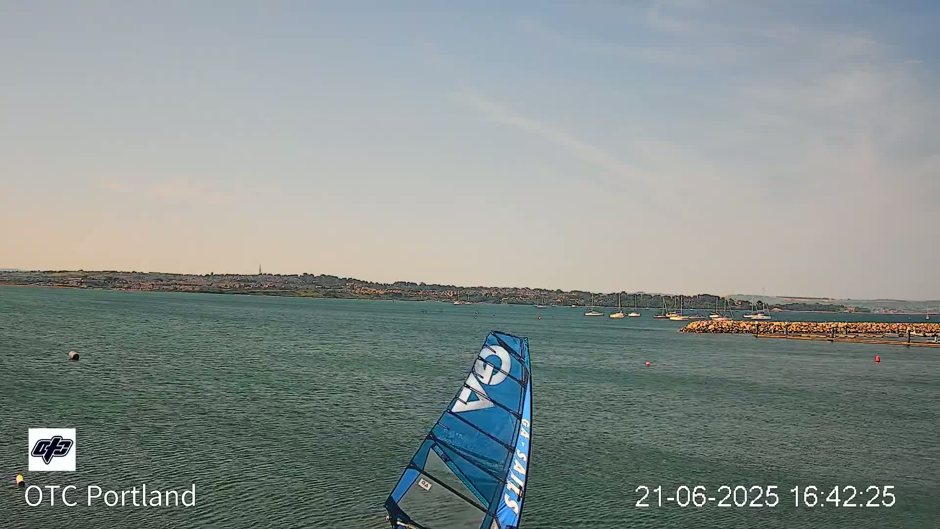 A windsurfer's sail is visible in the foreground of a calm body of water, with a distant shoreline and several boats under a mostly clear sky.