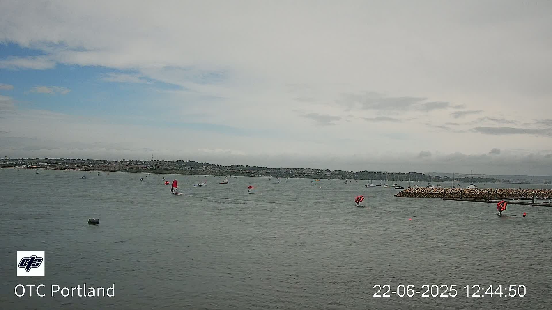 Numerous sailboats and windsurfers are scattered across a calm body of water under an overcast sky.