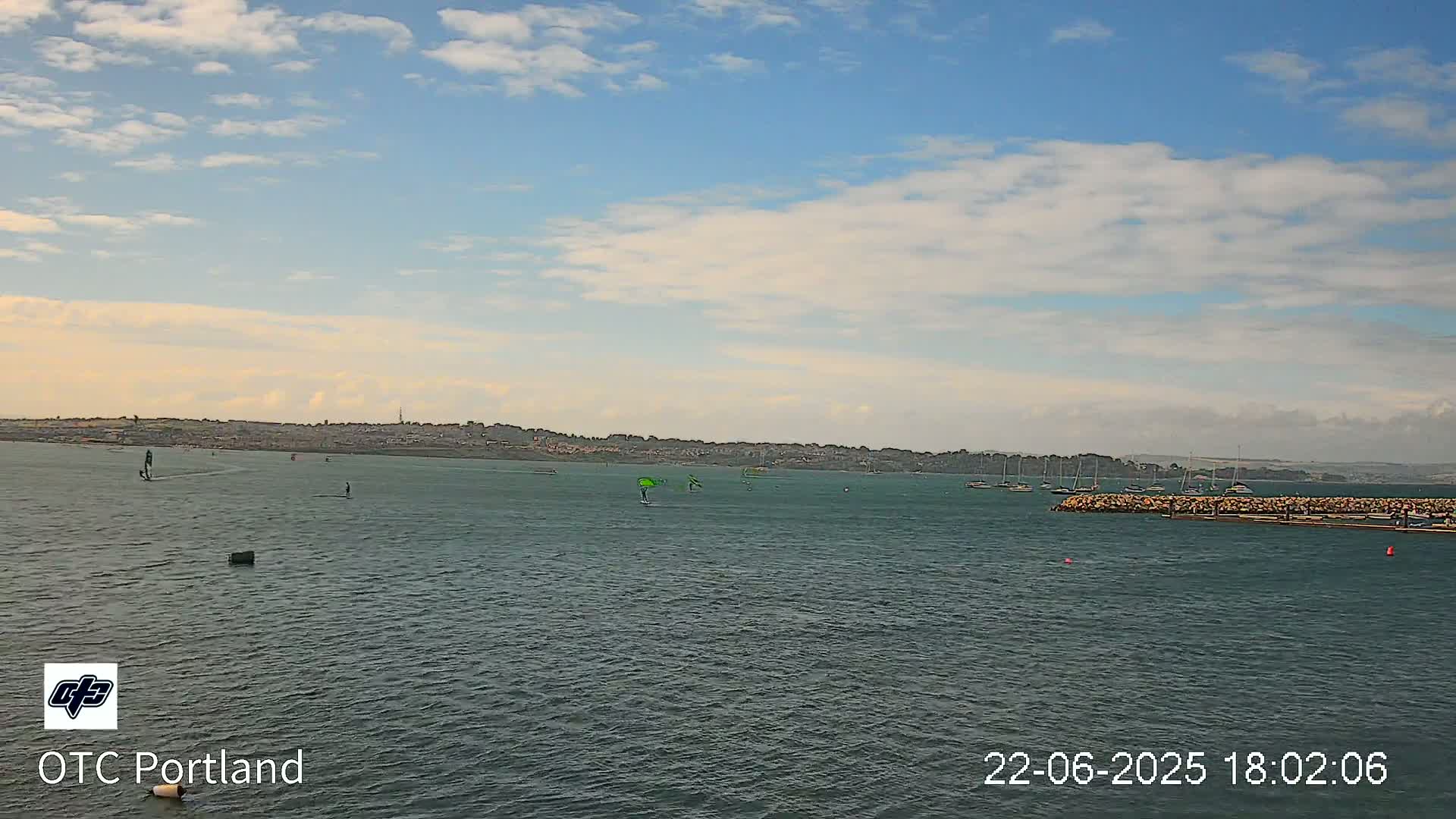 A calm body of water with several windsurfers and sailboats under a partly cloudy sky.