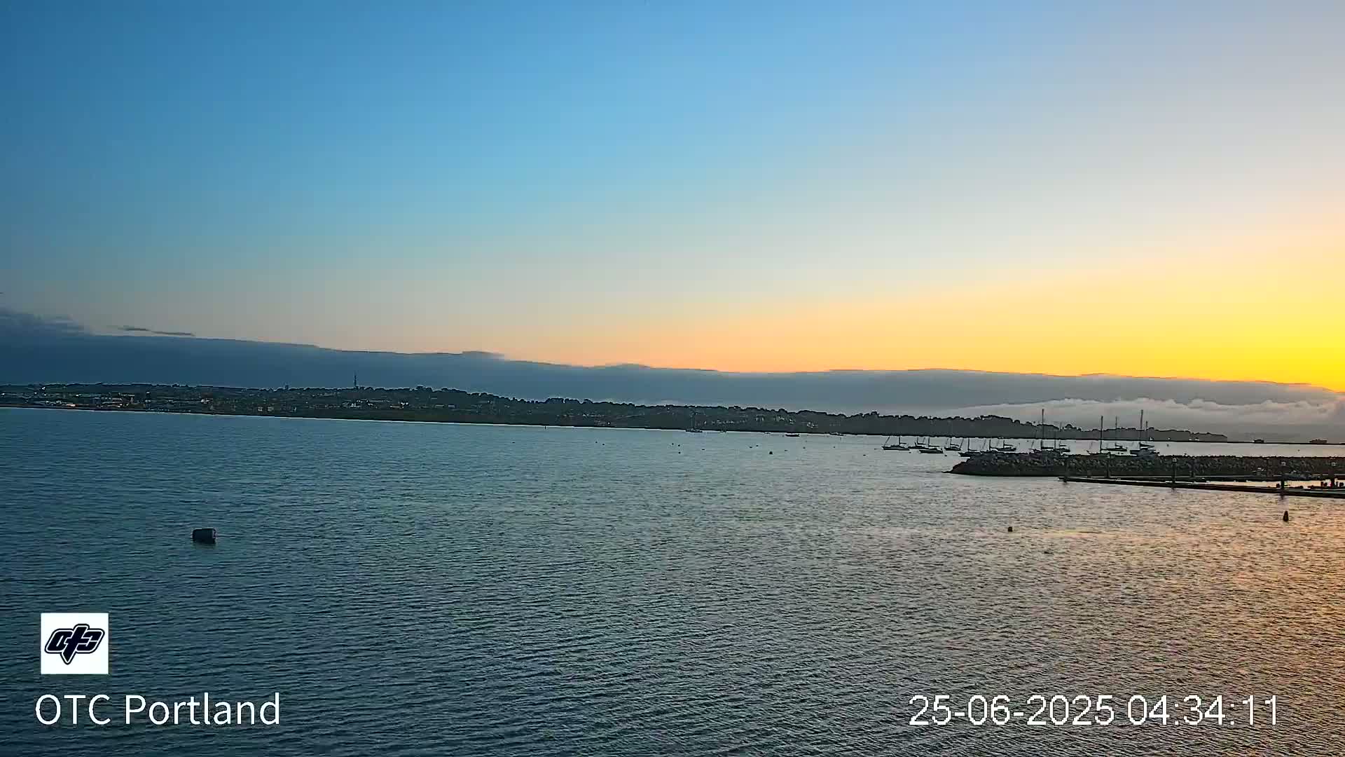 A calm body of water shows a distant shoreline at sunset, with a layer of fog or low clouds over the land.