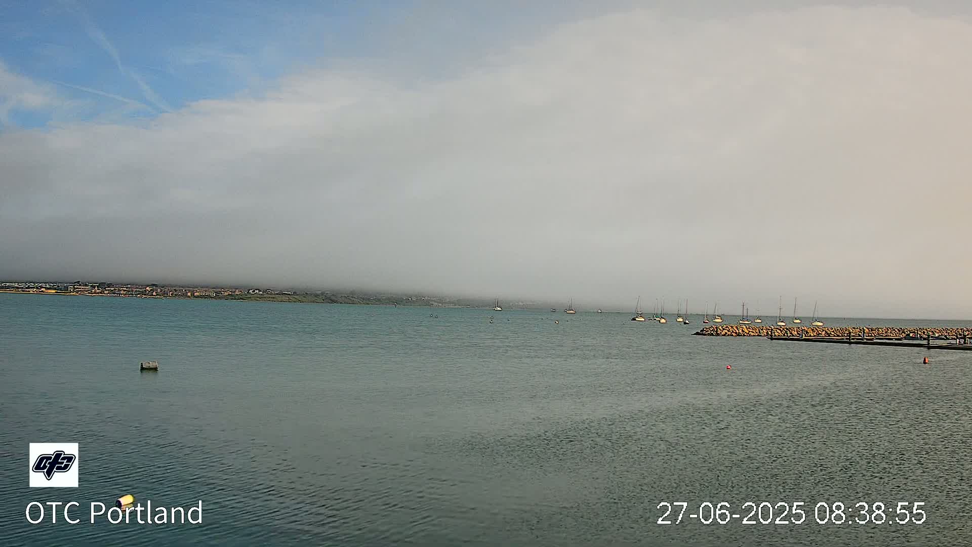 A calm body of water with several sailboats is visible, with a low-lying fog obscuring a distant shoreline under a mostly cloudy sky.