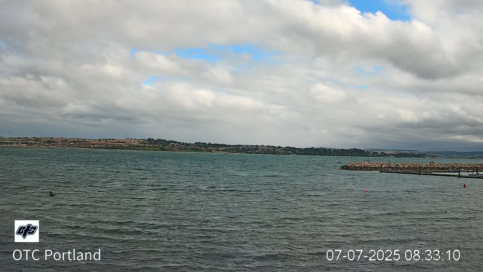 A body of water with a few small boats is seen under a mostly cloudy sky, with a distant shoreline visible.