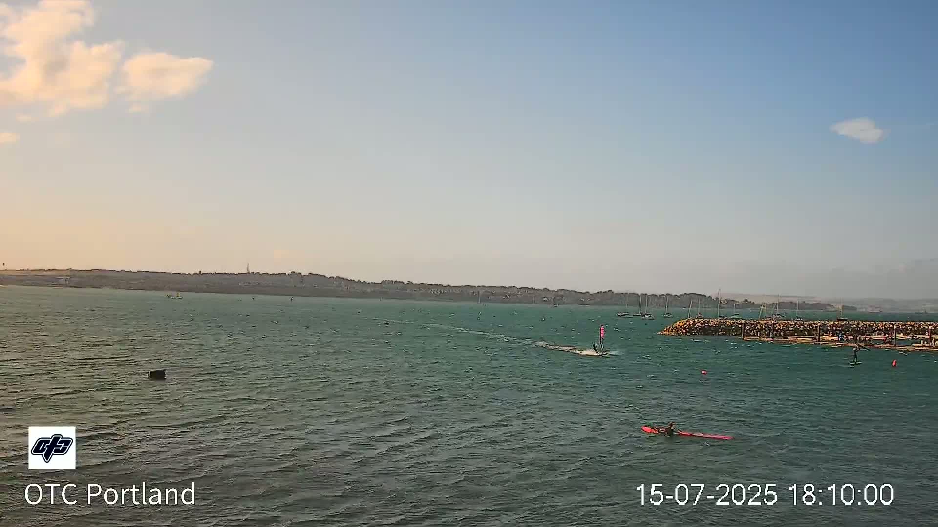 A calm, somewhat overcast day shows a body of water with several people windsurfing near a stone jetty and a distant shoreline.
